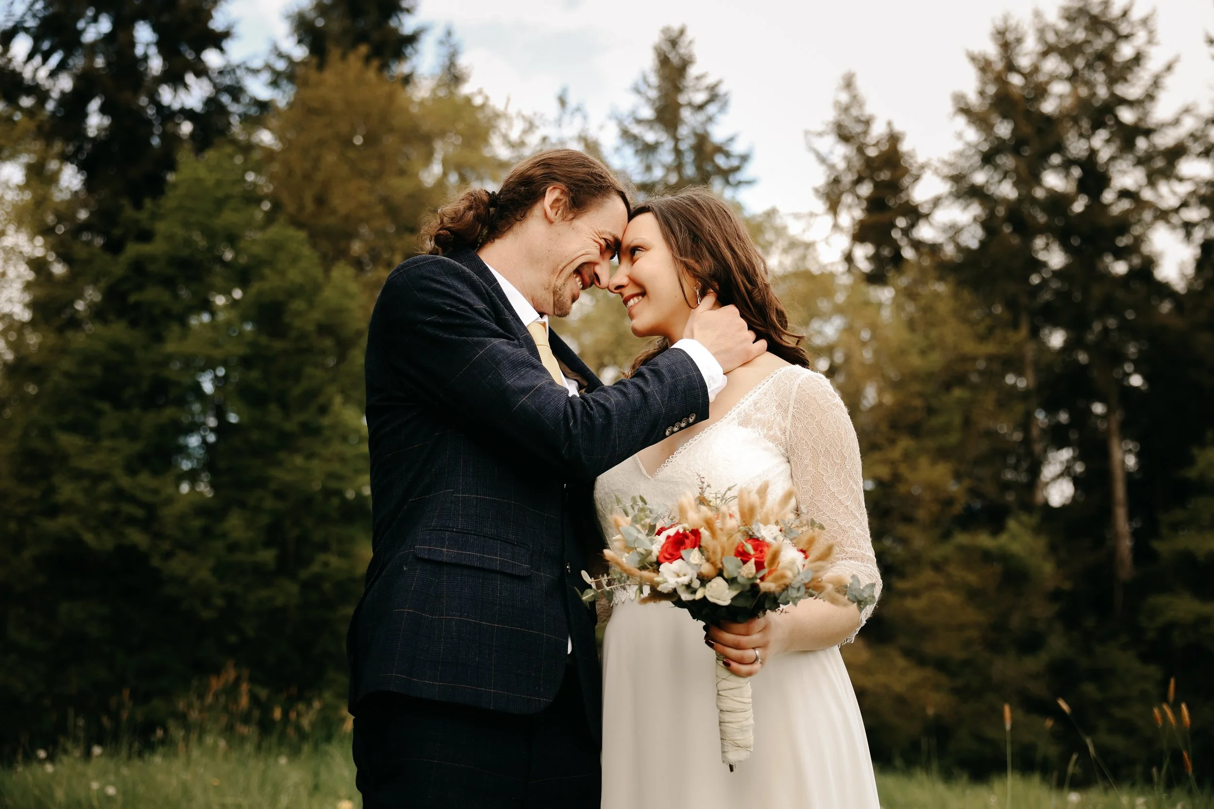 Un couple de mariage se regarde tendrement, avec leurs fronts touchant, dans un cadre naturel avec des arbres en arrière-plan. La femme porte une robe blanche et tient un bouquet de fleurs, tandis que l'homme porte un costume sombre. L'ambiance est j