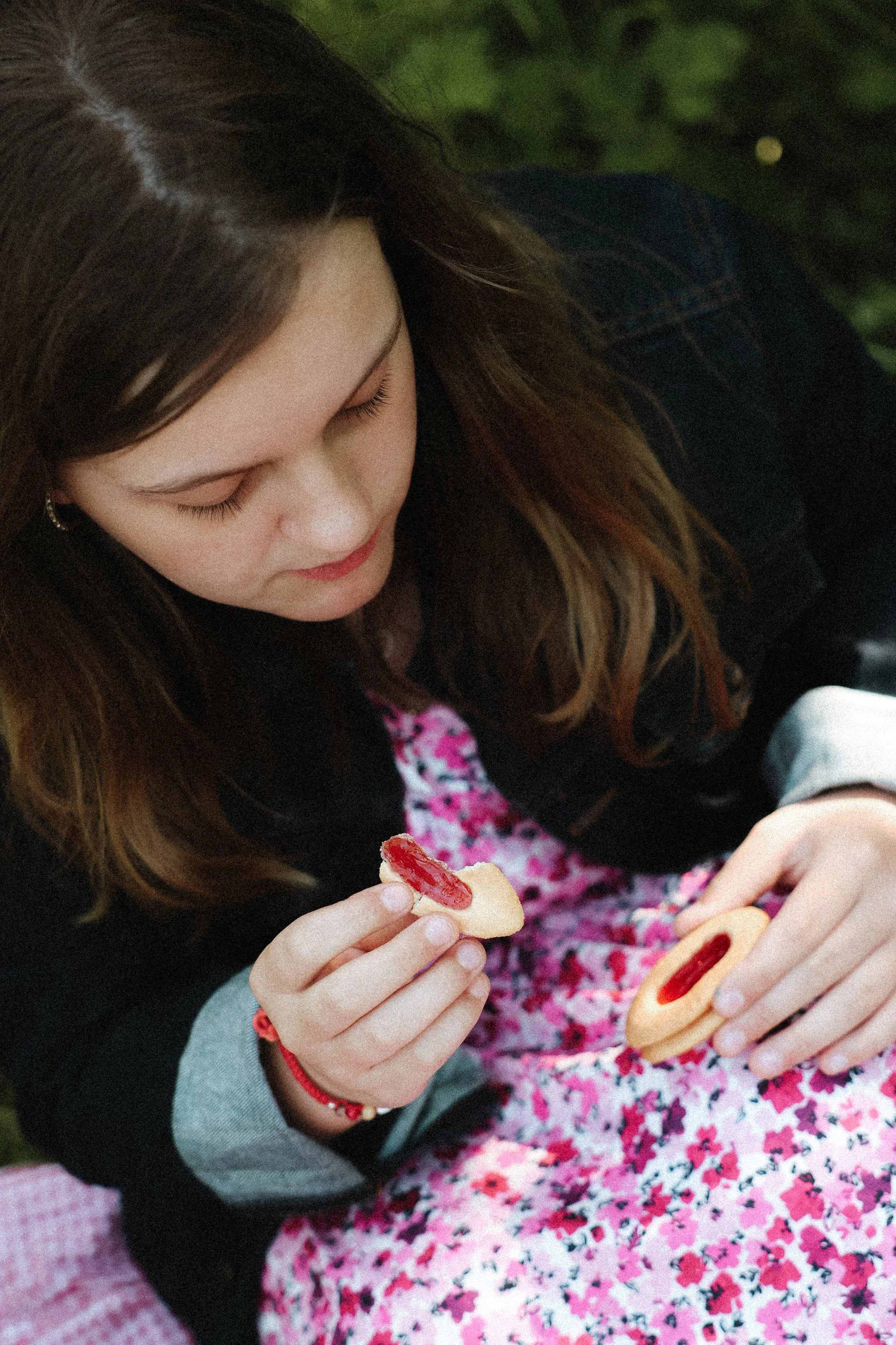 Une jeune fille avec des cheveux longs et bruns tient un biscuit avec de la confiture de fraise, elle est assise sur un vêtement rose à motifs floraux.