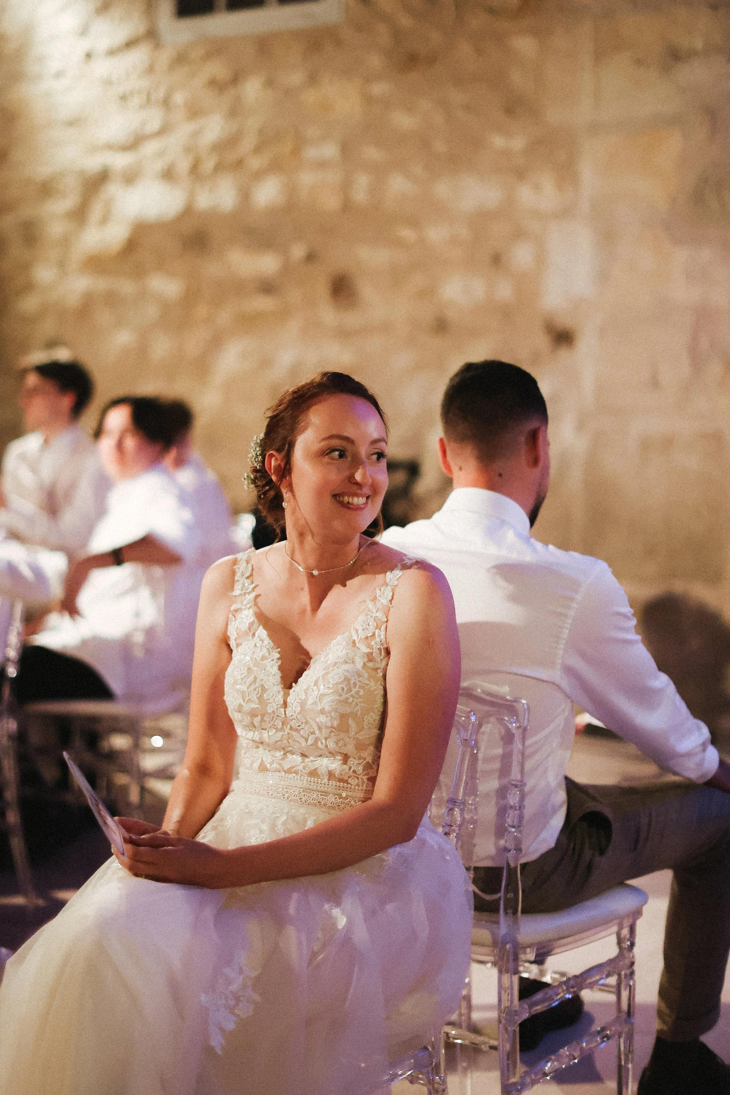 Une femme en robe de mariage assise dans une salle, souriant et tenant une photo, avec des autres personnes en arrière-plan lors d'une célébration ou d'un mariage.
