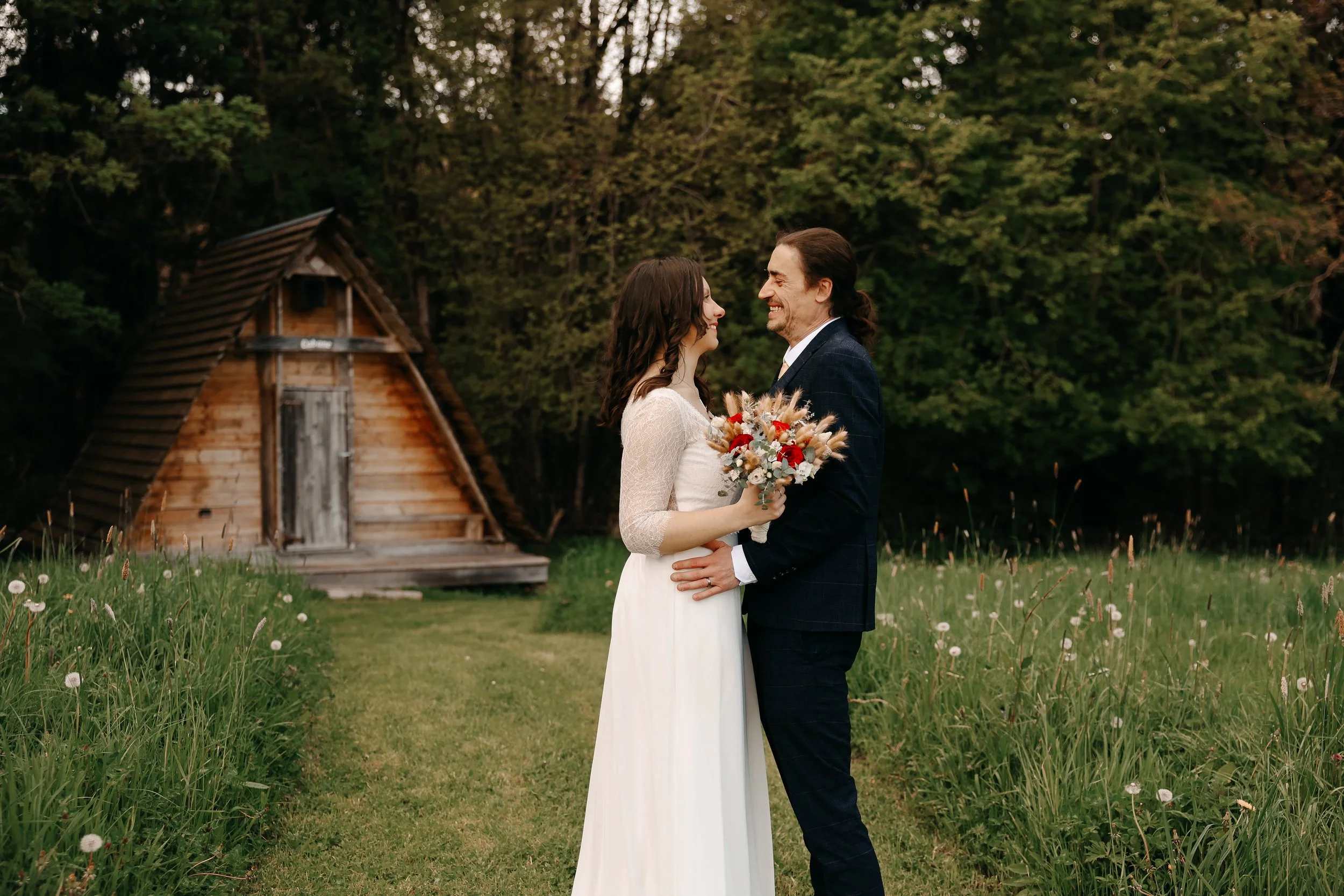 Un couple en mariage regarde chacun l'autre en souriant, debout dans un champ avec un bouquet de fleurs, avec une petite cabane en bois et des arbres en arrière-plan.