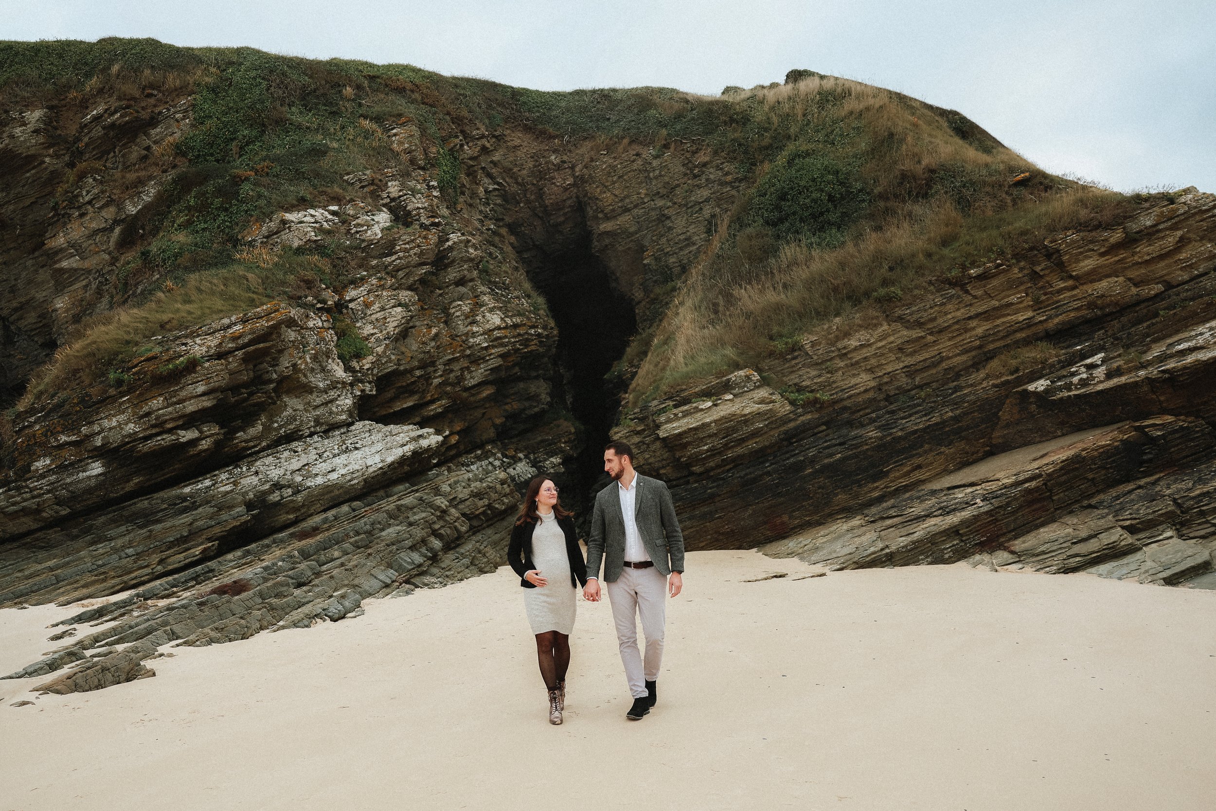 Un couple marche main dans la main sur une plage avec des falaises rocheuses en arrière-plan.