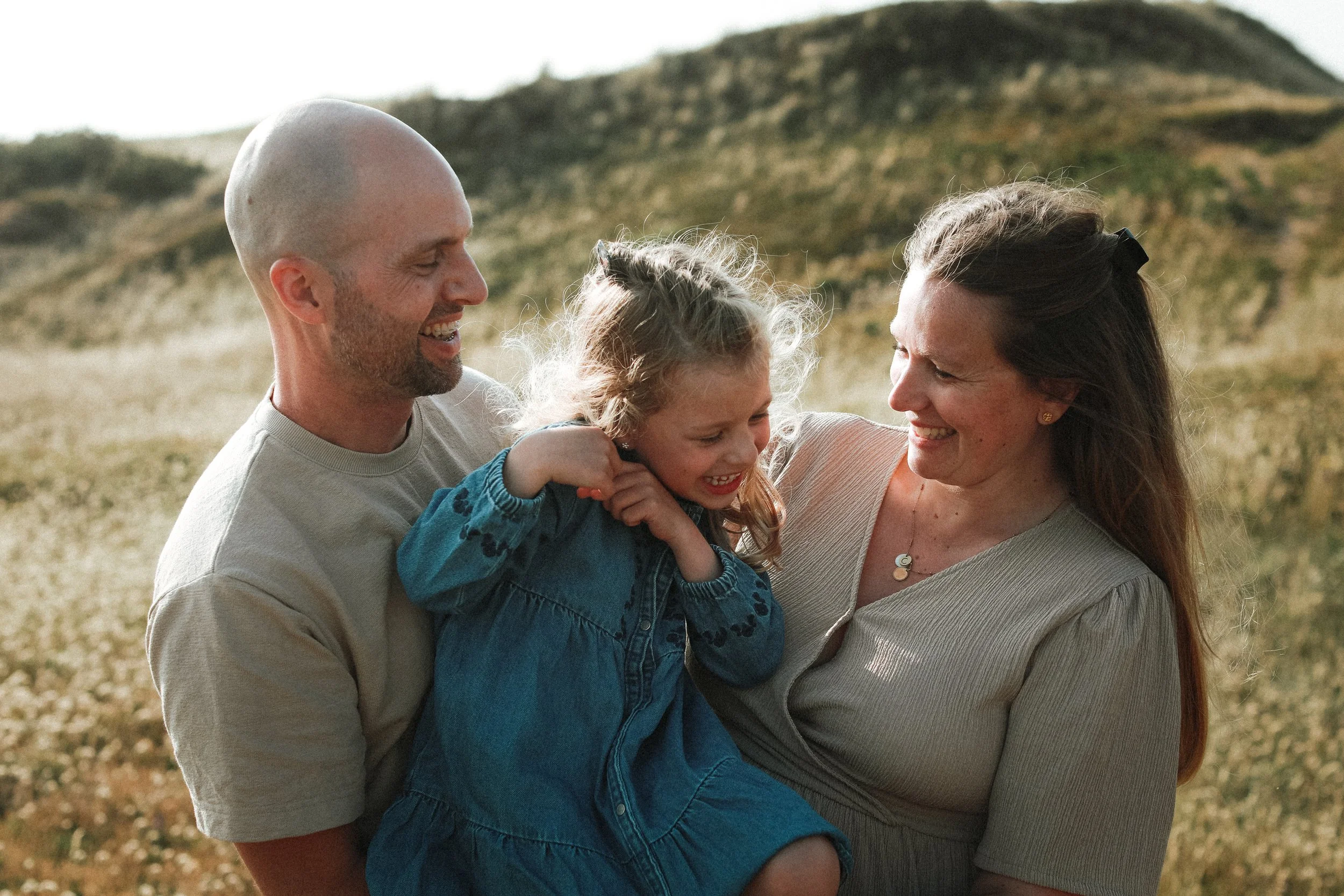 Une famille de trois personnes, un père, une mère et une fille, souriants, en plein air dans un champ avec des collines en arrière-plan. La fille est assise sur la mère, et le père tient la fille dans ses bras, tous rient ensemble.