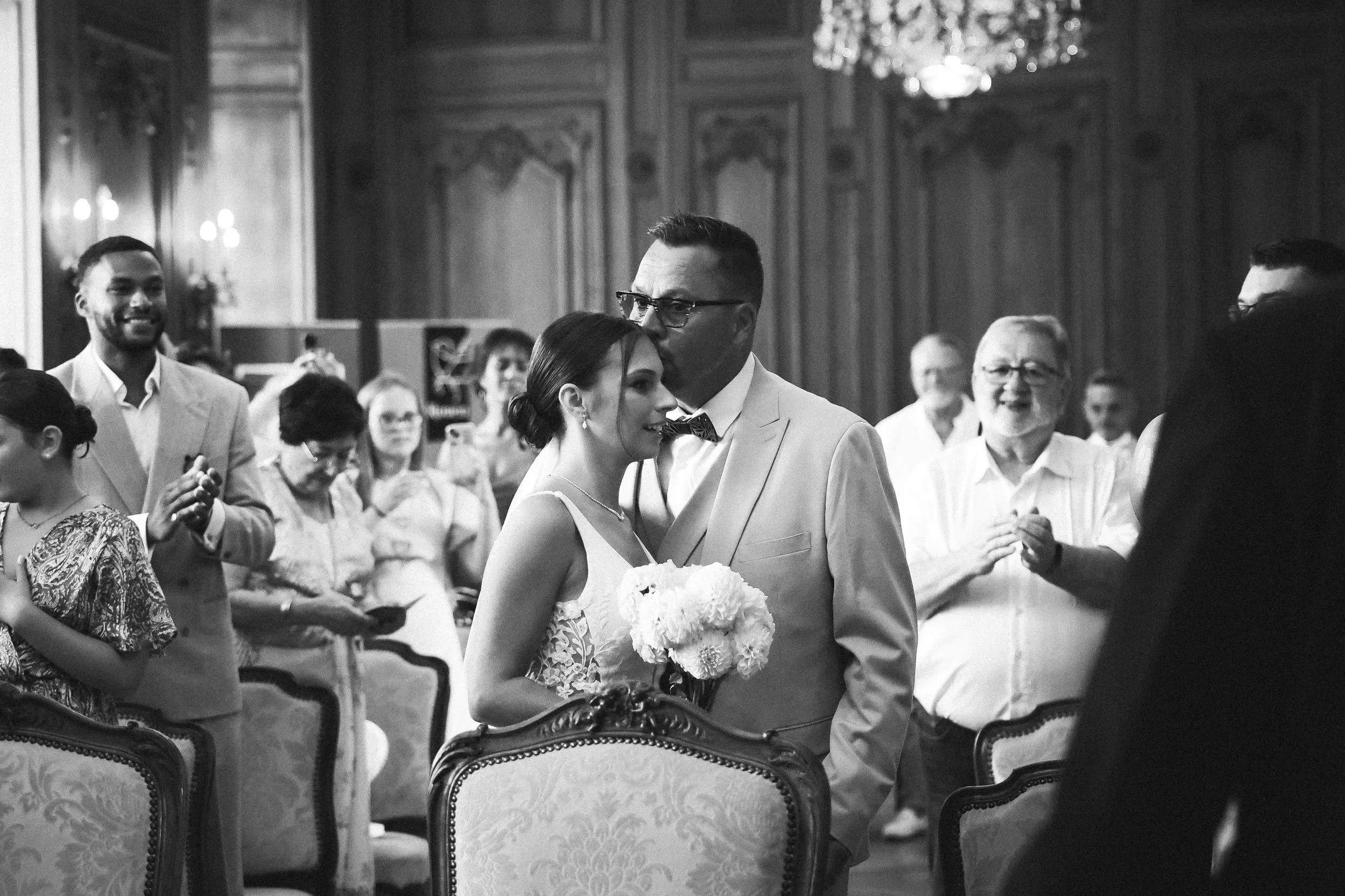 Un couple de mariés lors de leur cérémonie de mariage, entourés d'invités dans une salle élégante. La mariée tient un bouquet de fleurs et porte une robe blanche, tandis que le marié porte un costume clair avec un nœud papillon. Tout le monde semble 