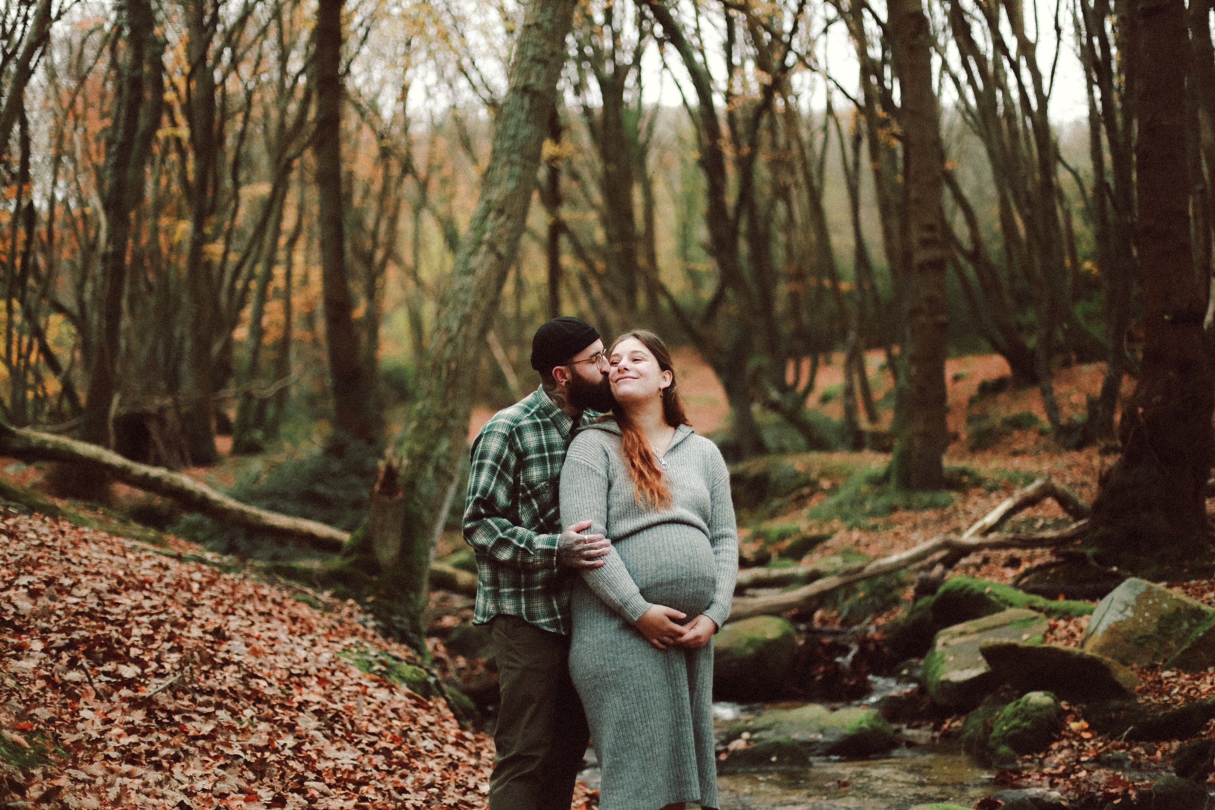 Un couple, dont une femme enceinte, dans une forêt aux arbres aux feuilles d'automne, face à un petit ruisseau.