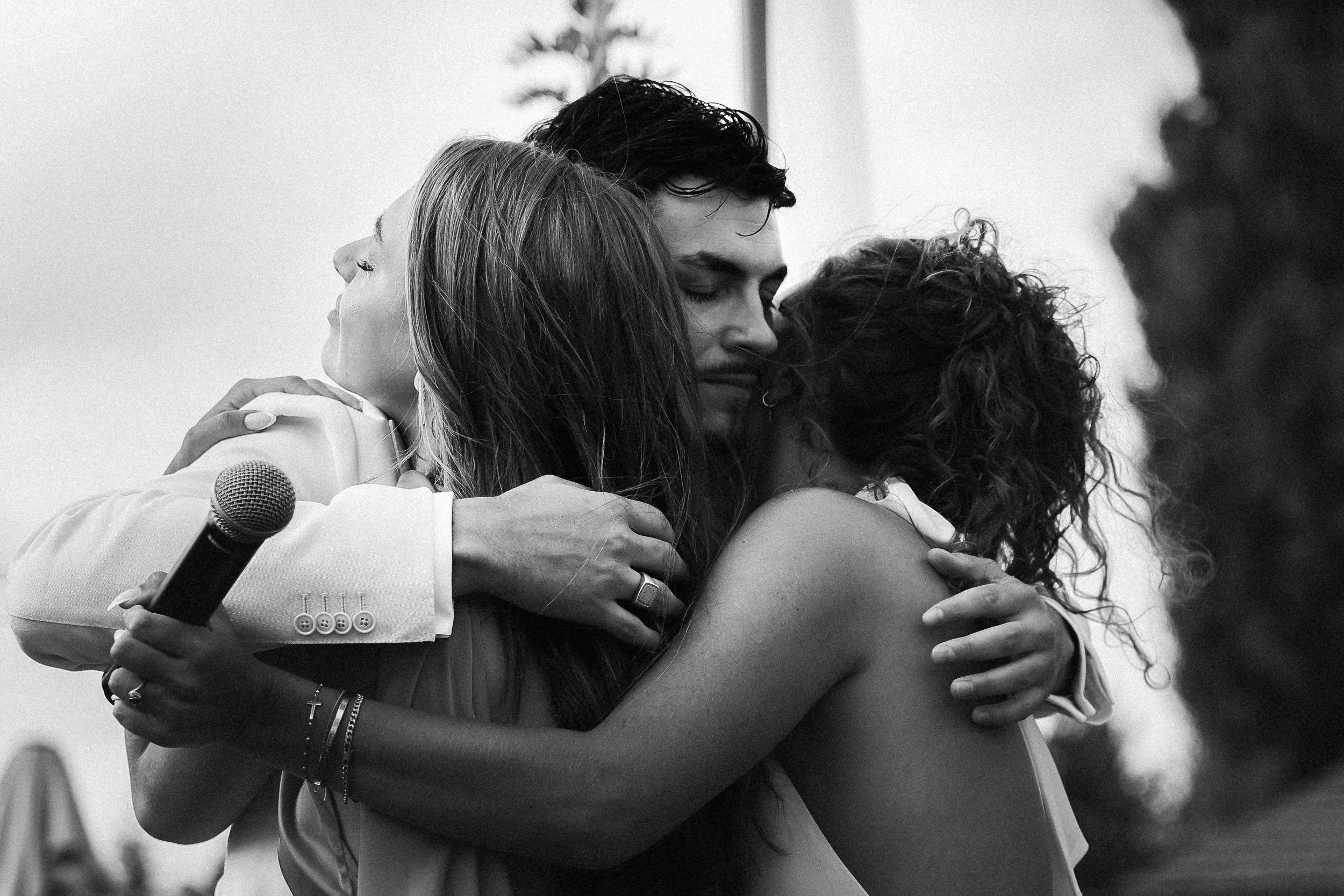 Trois personnes s'étreignent dans une accolade chaleureuse, avec un micro en main, lors d'un événement extérieur, en noir et blanc.