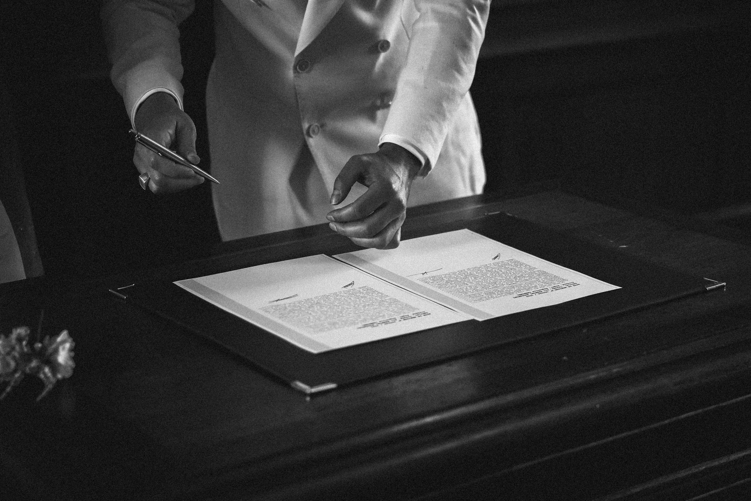 Un document officiel étalé sur une table, la main d'une personne en costume dessus, dans une ambiance sombre.