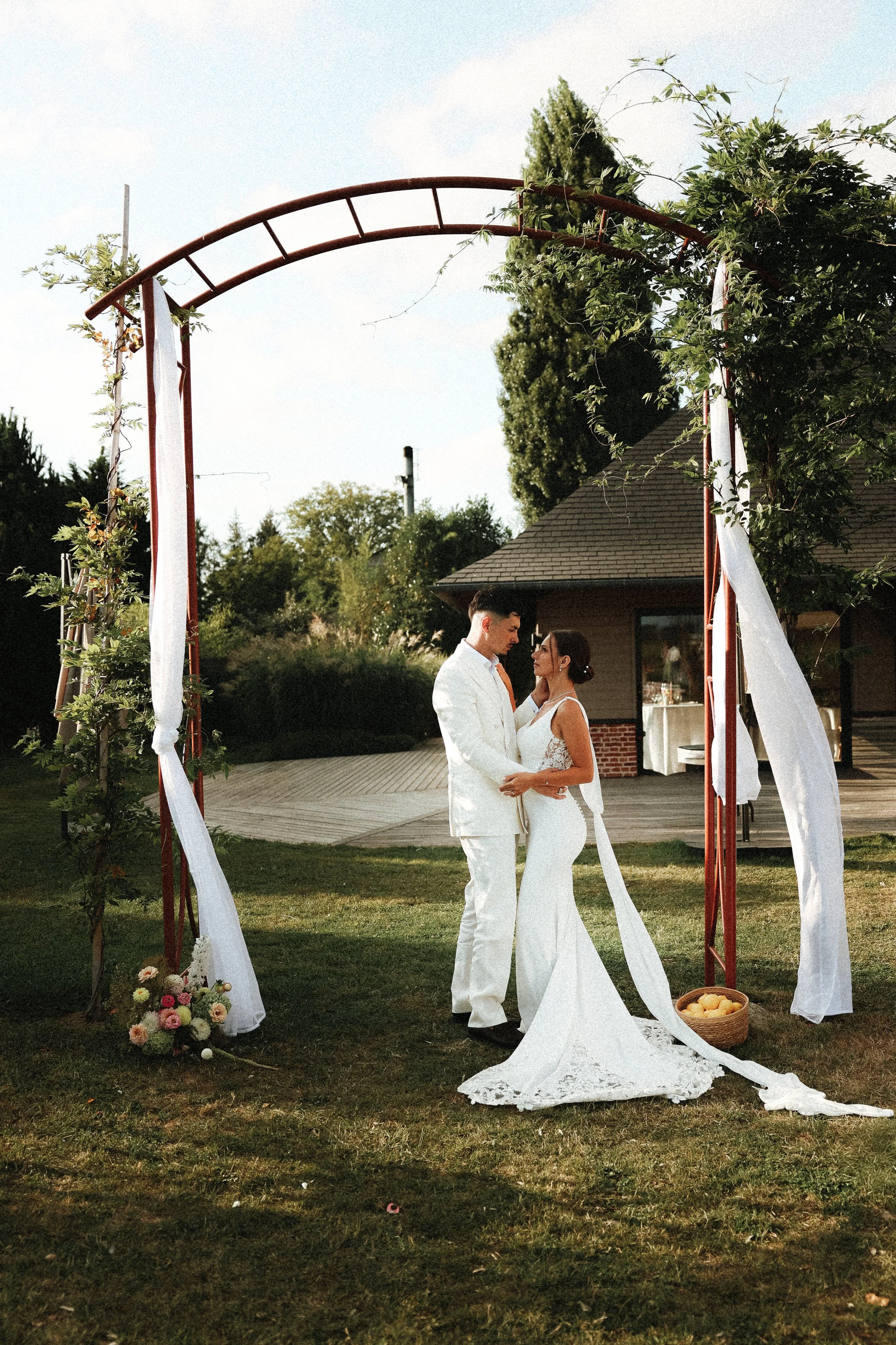 un couple de mariés lors de leur mariage en plein air sous une arche décorée de tissus blancs et de verdure, avec une maison en arrière-plan