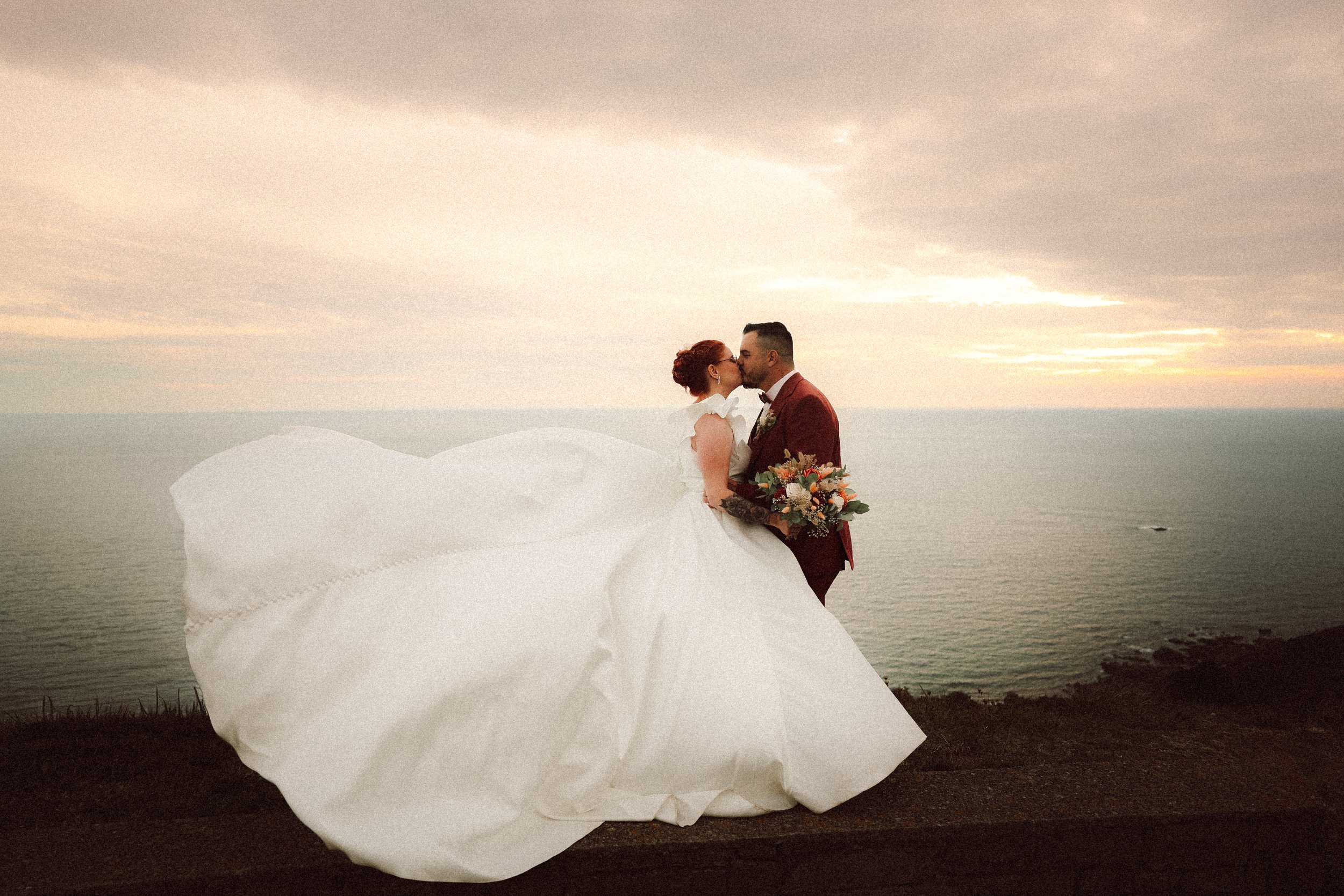Un couple de mariés s'embrassant lors d'une séance photo au coucher du soleil au bord de la mer, avec la mariée en robe blanche et le marié en costume rouge foncé, tenant un bouquet de fleurs.