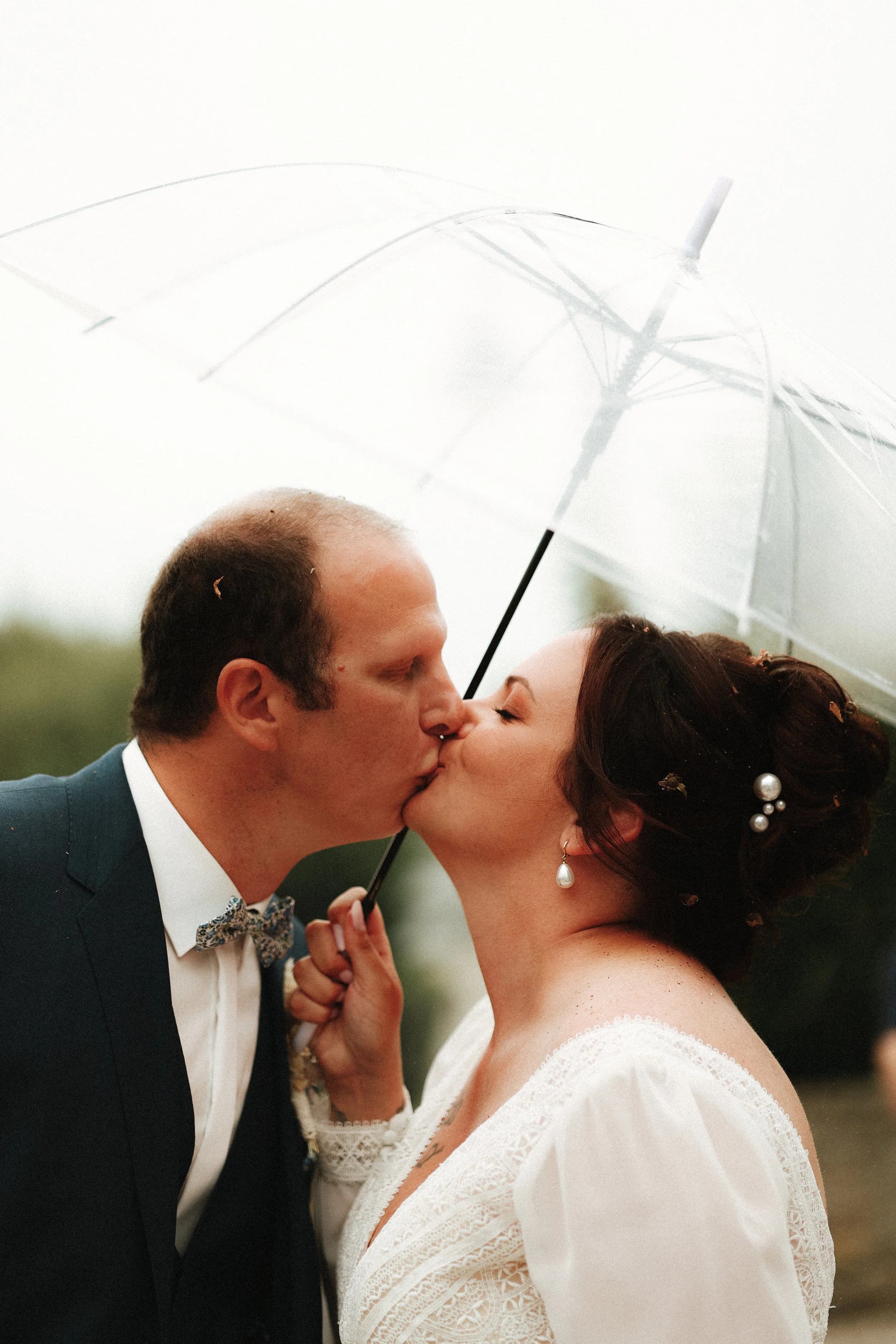 Un couple de mariés s'embrasse sous un parapluie lors d'une séance photo de mariage, plein air, fond flou vert.