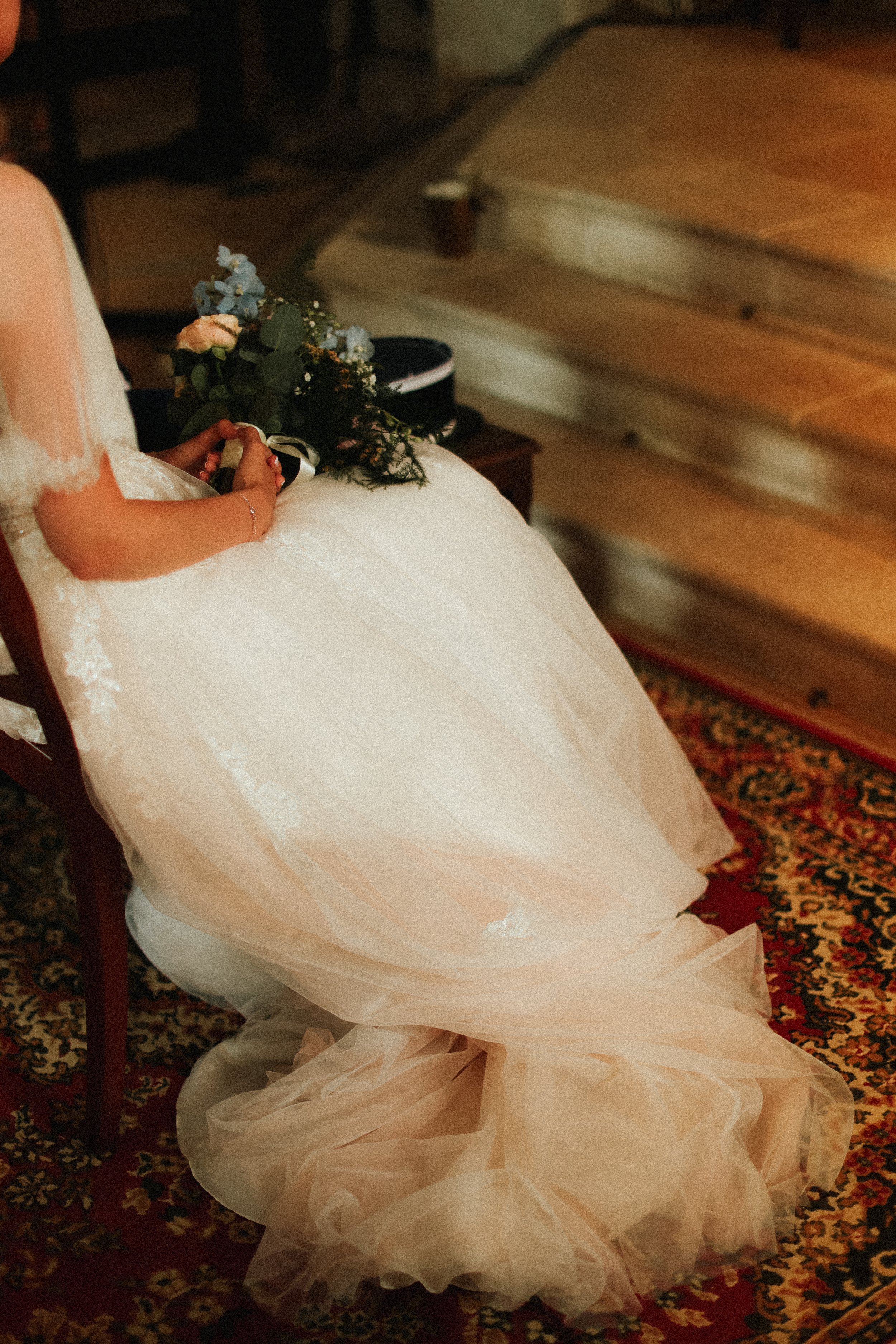 Une femme en robe de mariage tenant un bouquet de fleurs, assise sur une chaise, dans une pièce chauffée, avec un lit en bois en arrière-plan.