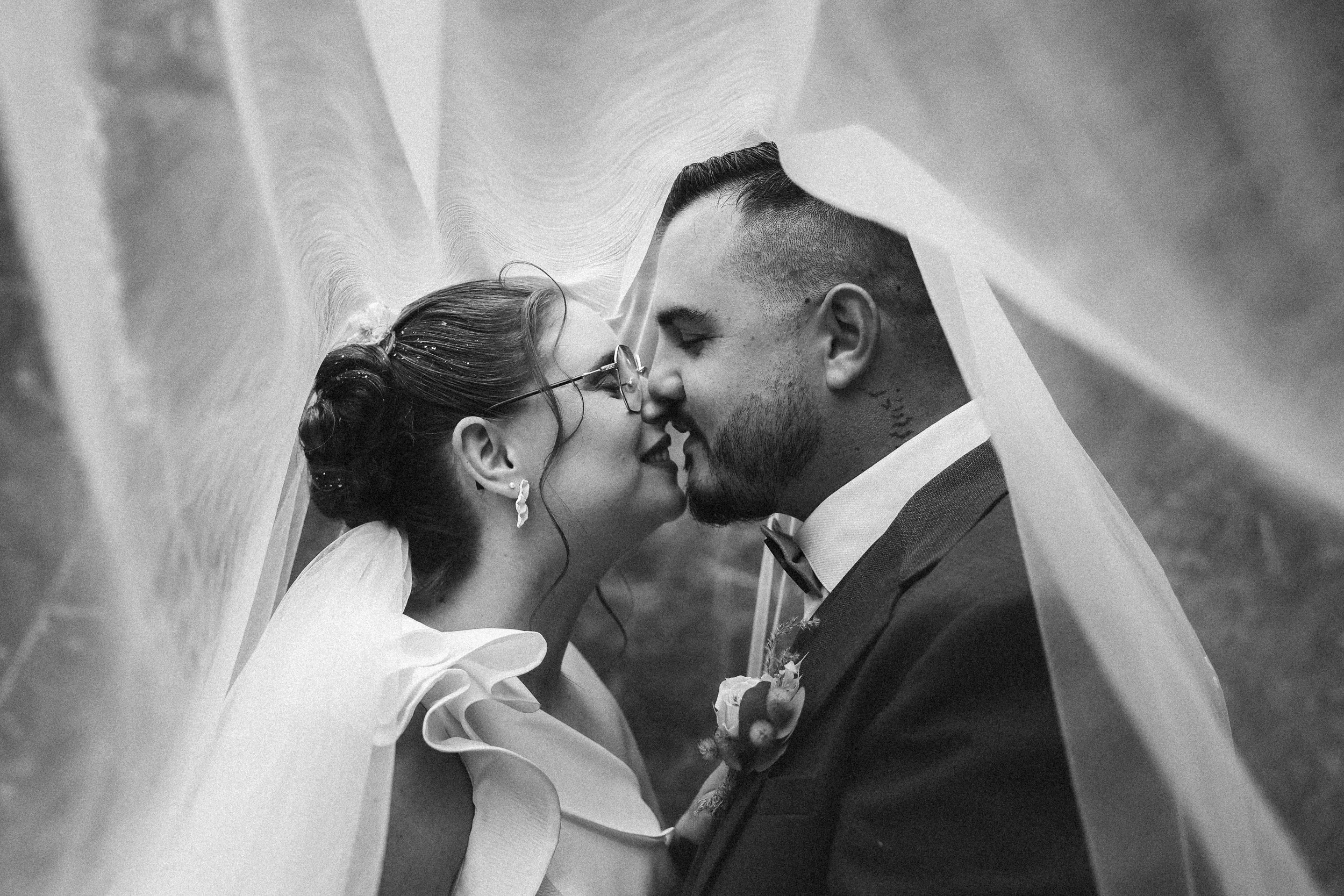 Un couple de mariés s'embrassant sous un voile lors de leur mariage, en noir et blanc.