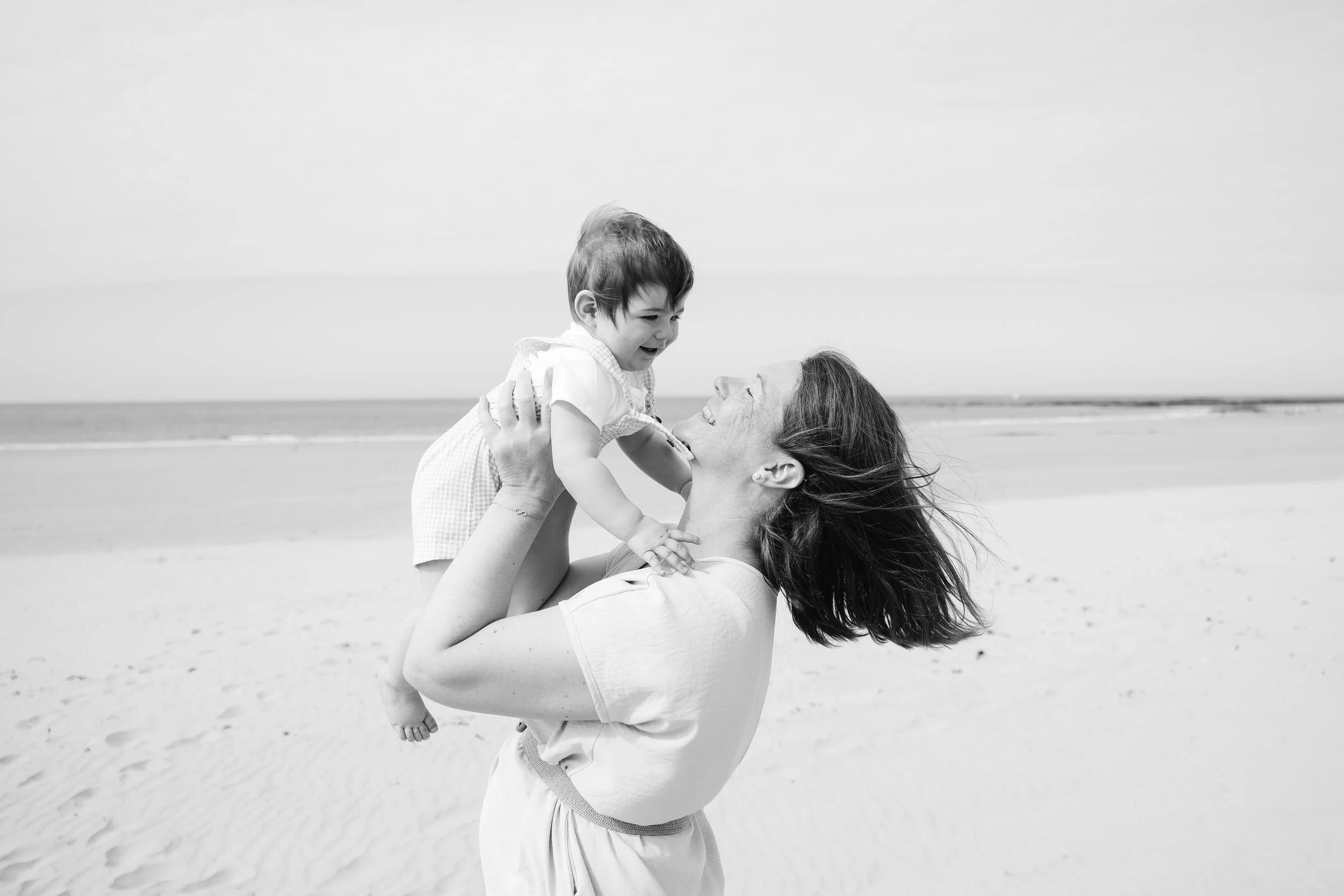 Une femme souriante tient un jeune enfant à hauteur de ses épaules sur la plage, tous deux semblant heureux. La scène est en noir et blanc, avec la mer en arrière-plan.