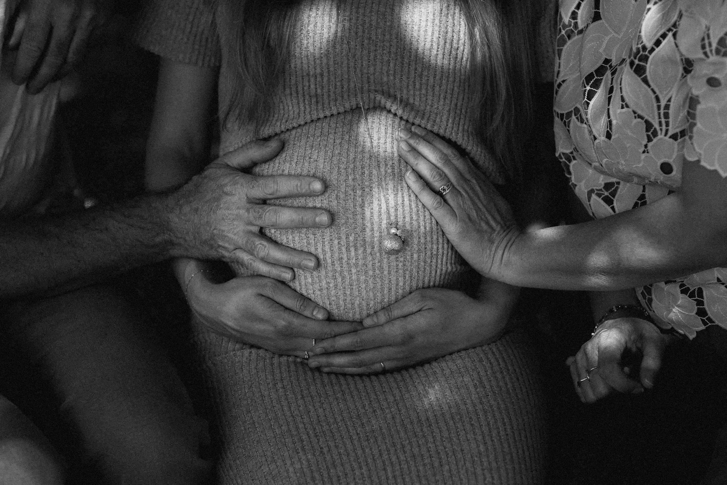 Une femme enceinte, vêtue d'un pull en maille, est entourée de plusieurs mains posées sur son ventre, symbolisant l'amour et la protection.