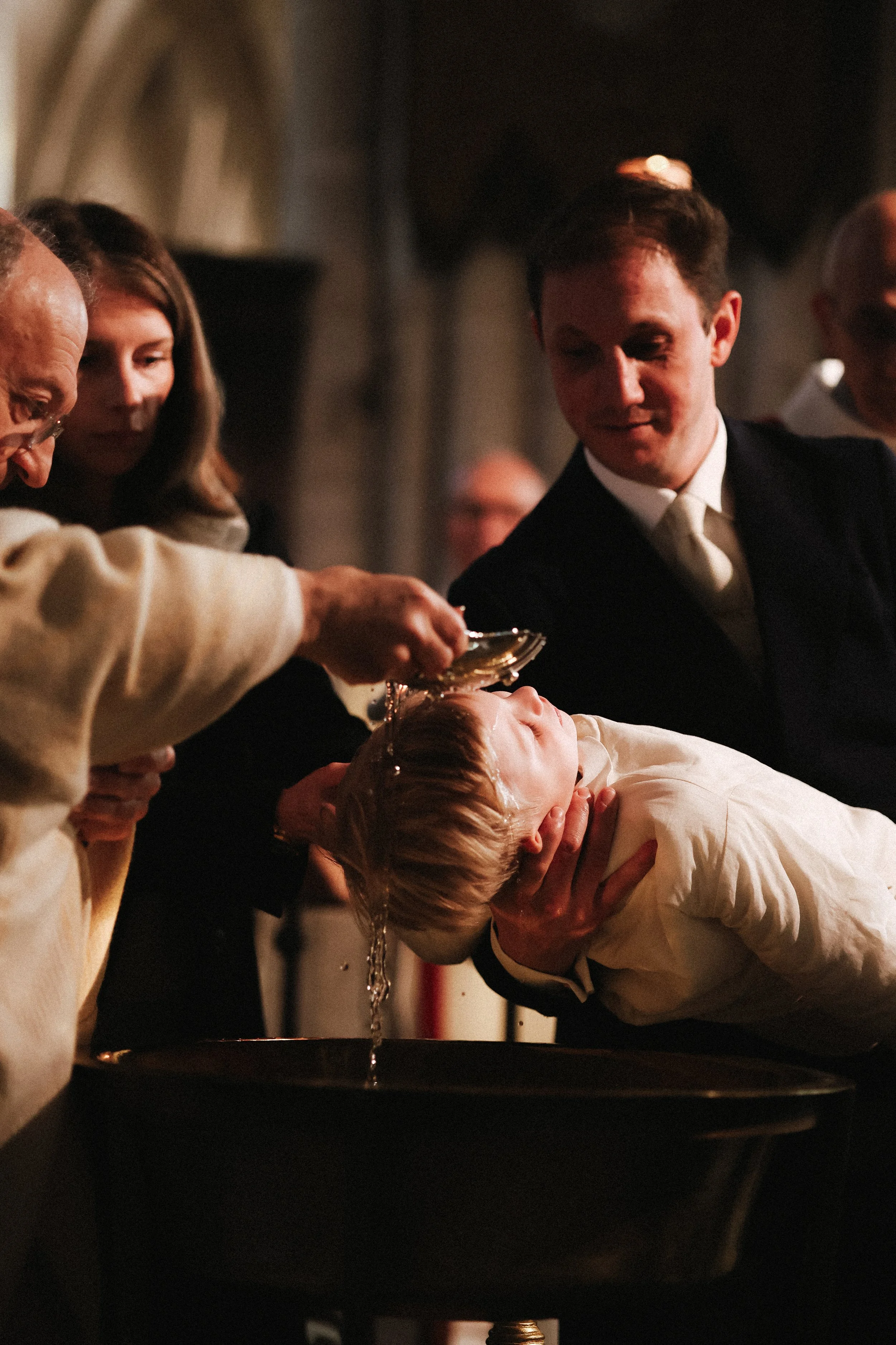 Un enfant reçoit le baptême dans une église, la personne en train de verser de l'eau sur sa tête est entourée d'autres personnes.