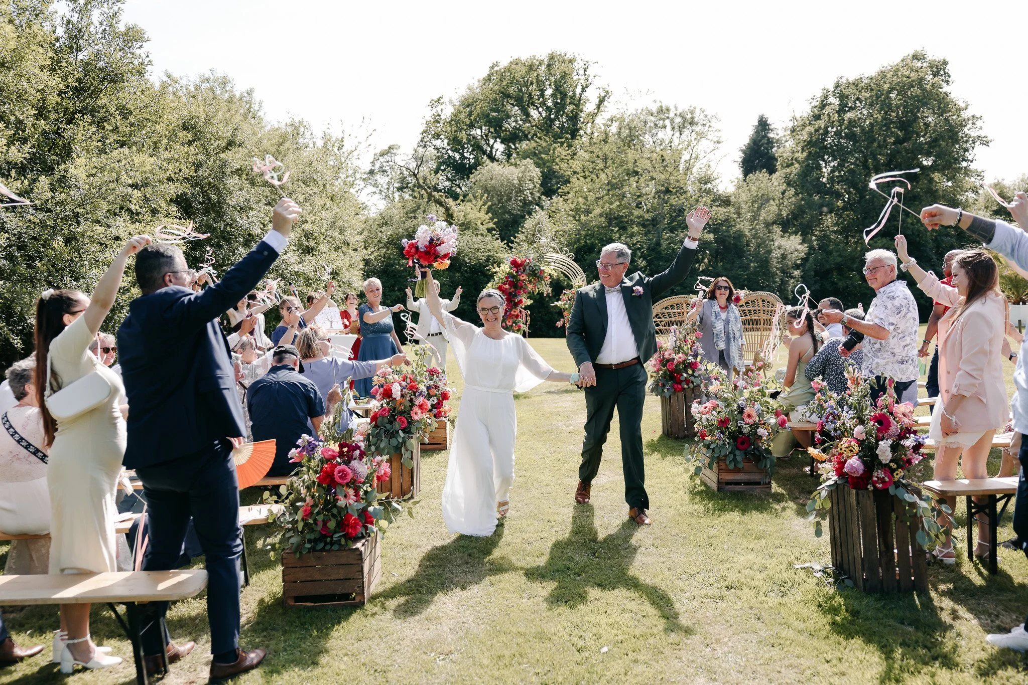 Un mariage en plein air avec un couple main dans la main marchant sous une arche fleurie, entouré de convives célébrant avec des confettis et des fleurs dans un jardin ensoleillé.