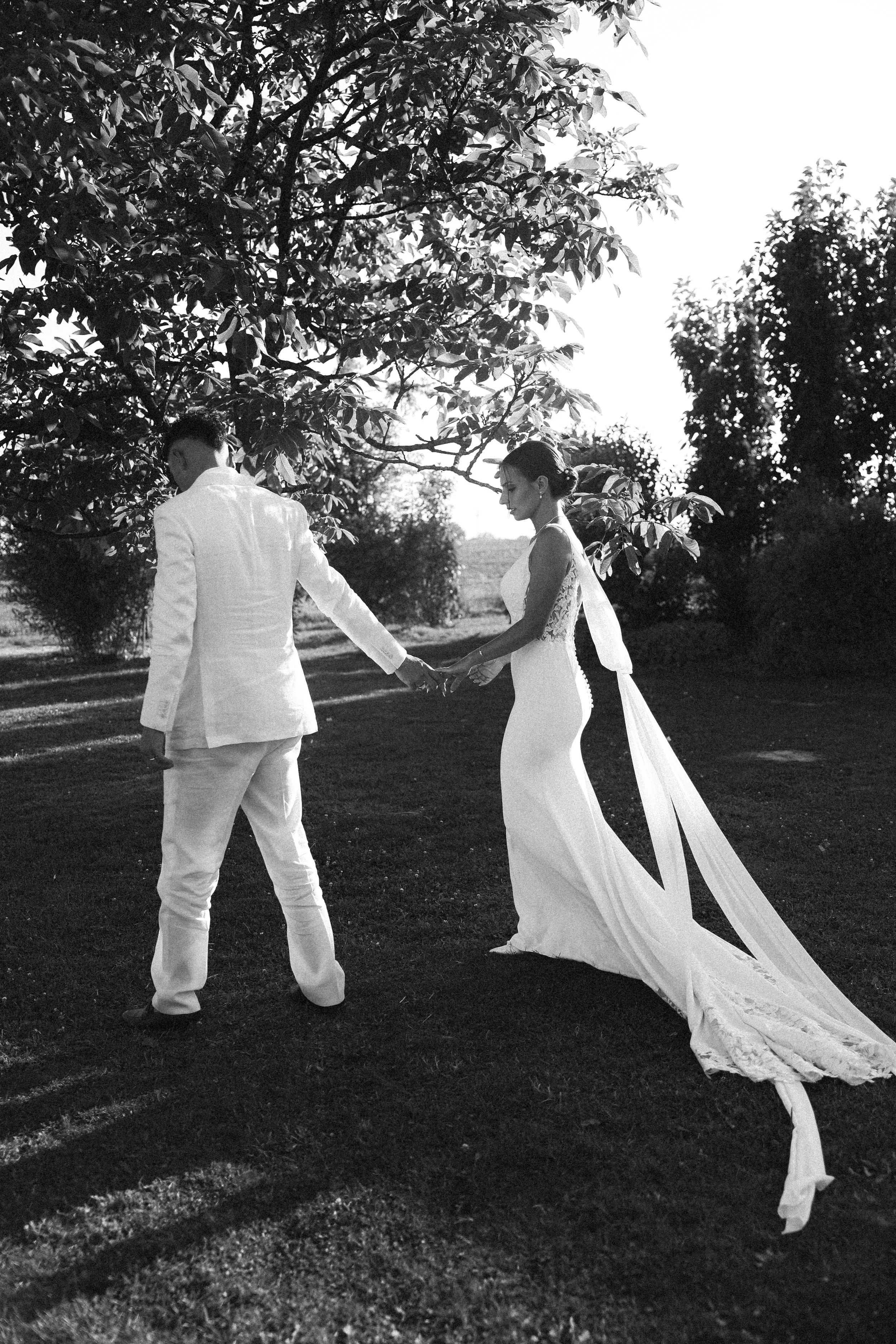 Un couple marié, une femme en robe de mariage et un homme en costume blanc, se tenant la main dans un espace extérieur, arbres en arrière-plan, la scène en noir et blanc.