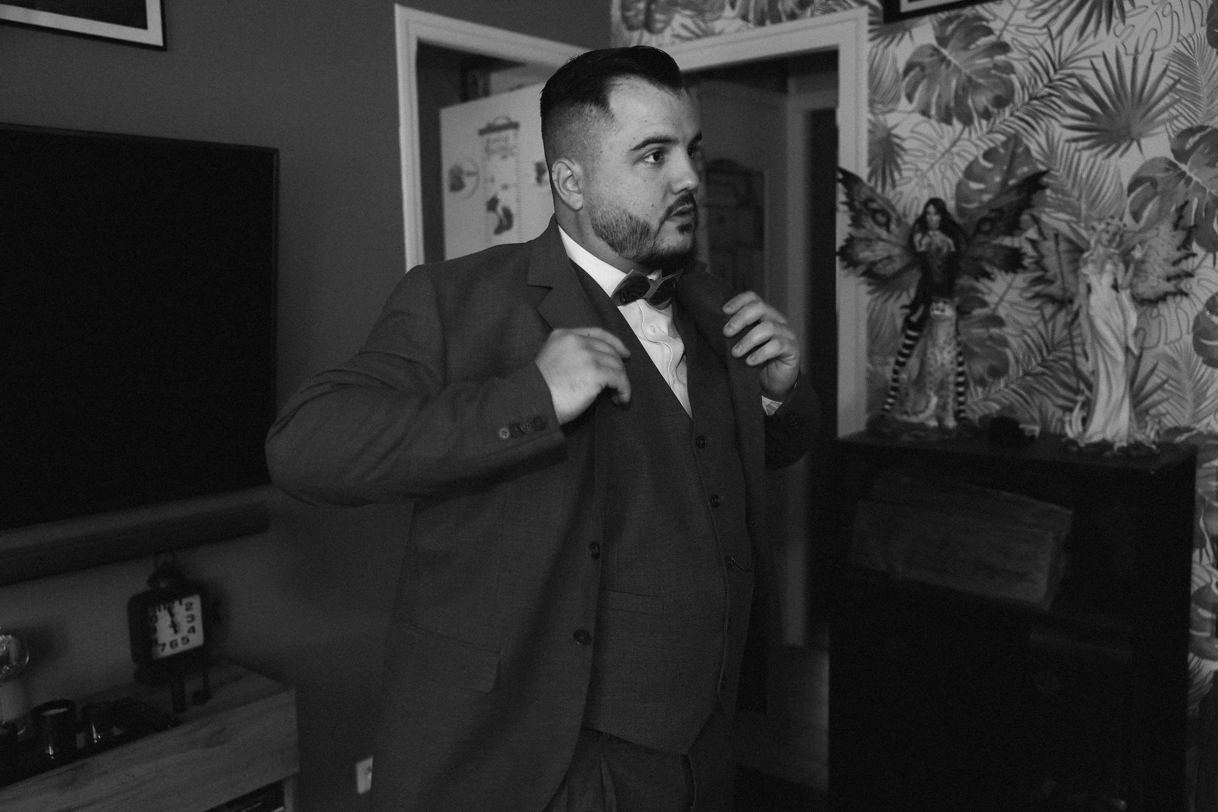 Un homme en costume ajustant sa veste dans une pièce décorée avec des motifs végétaux, en noir et blanc.