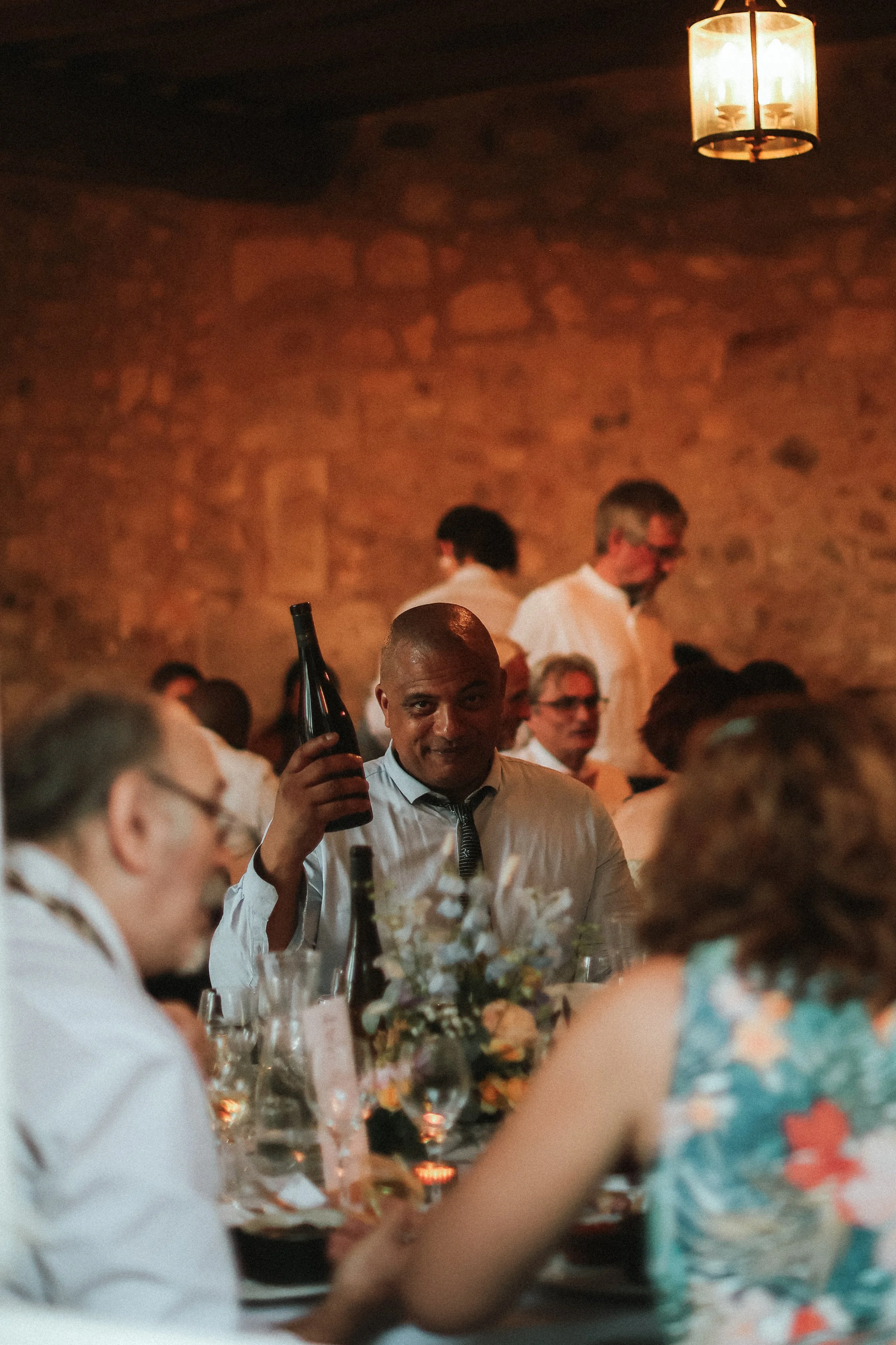 Groupe de personnes lors d'un dîner, une personne au premier plan porte une chemise blanche et soulève une bouteille de vin, ambiance chaleureuse avec un mur en pierre en arrière-plan.