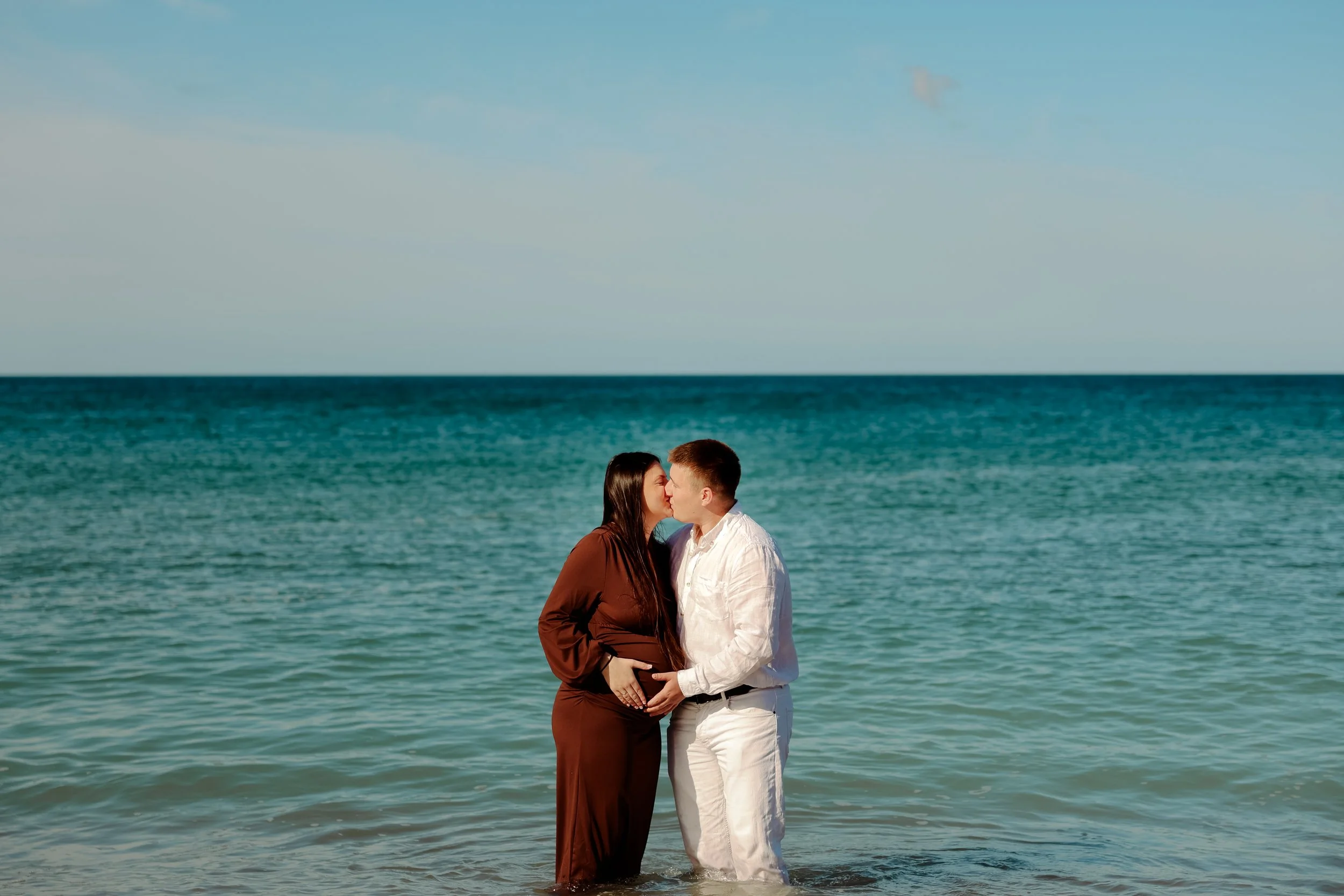 Un couple en tenues décontractées se tenant par la taille dans la mer, s'embrassant, avec la mer et le ciel en arrière-plan.