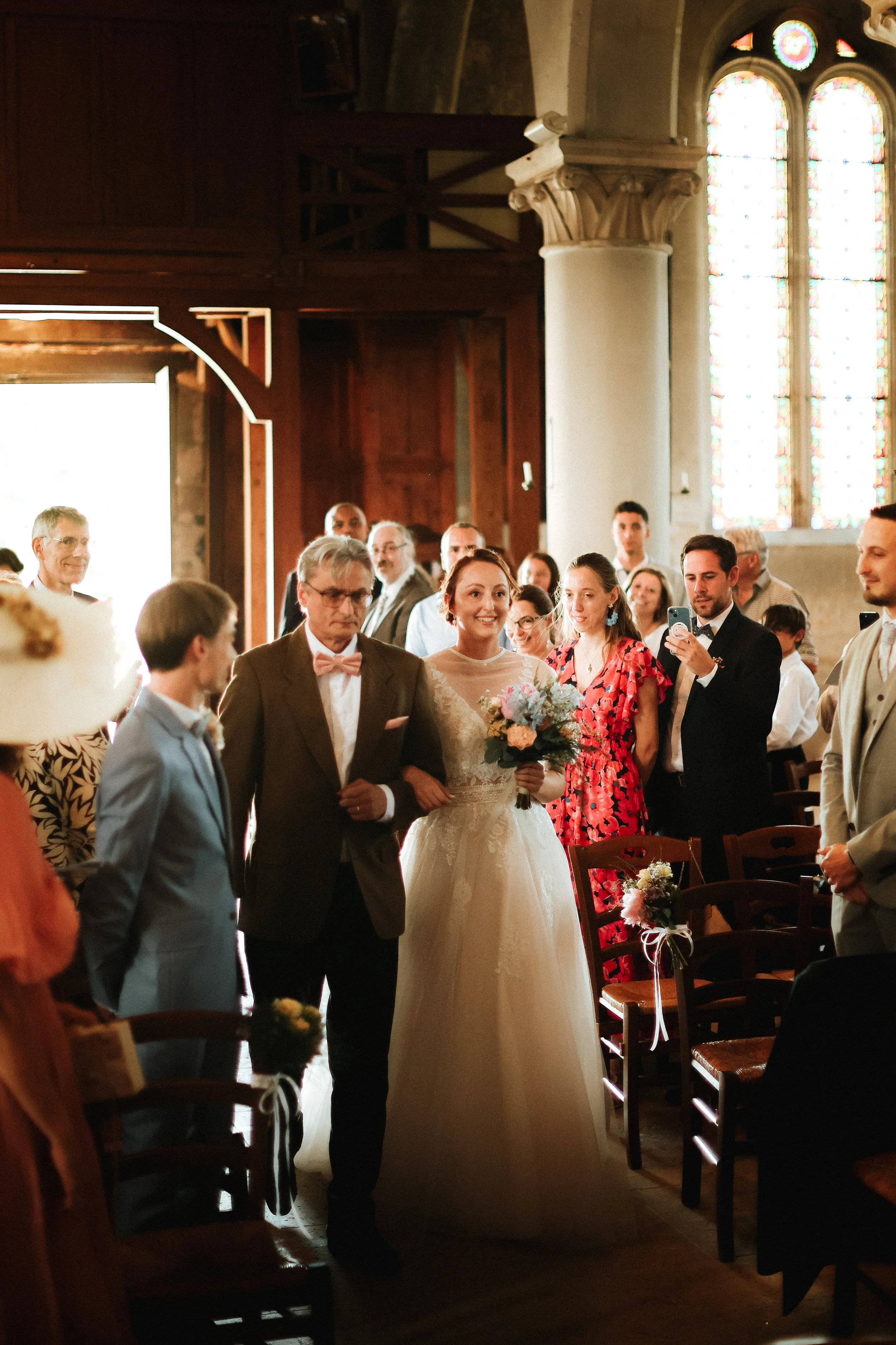 Une mariée marchant dans une église, accompagnée par un homme (probablement son père), entourée de personnes souriantes et en tenue élégante, pendant un mariage.