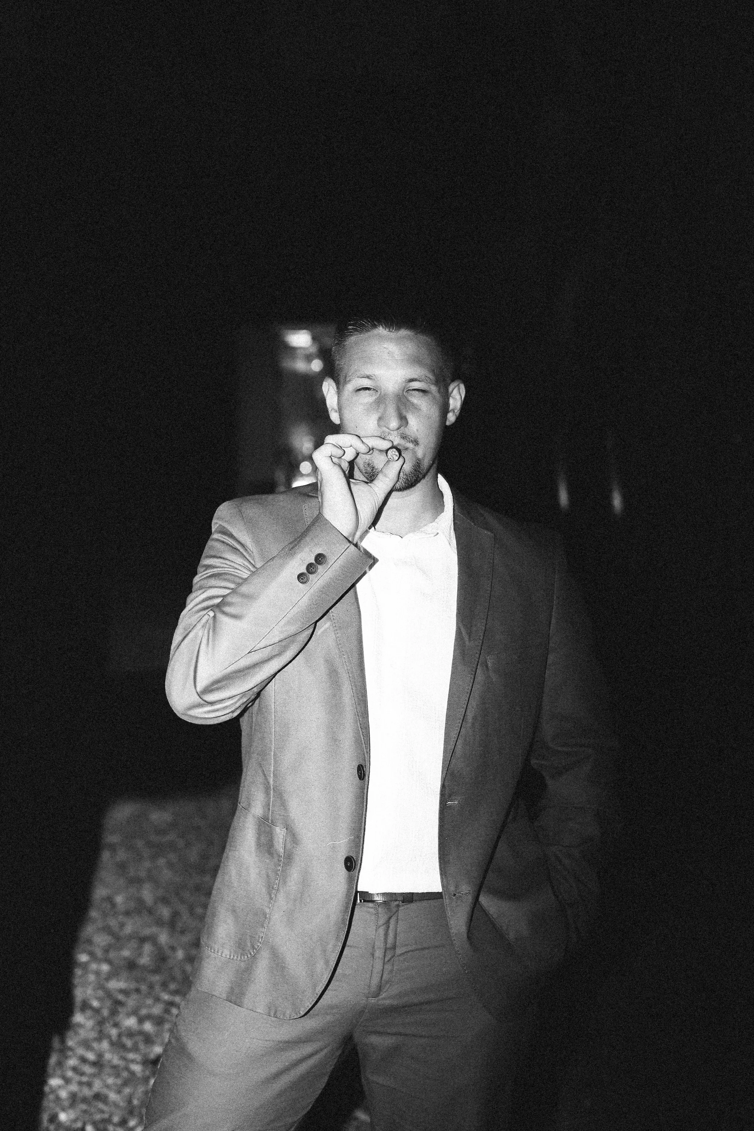 Un homme en costume, fumant une cigarette, portrait en noir et blanc dans un environnement sombre.