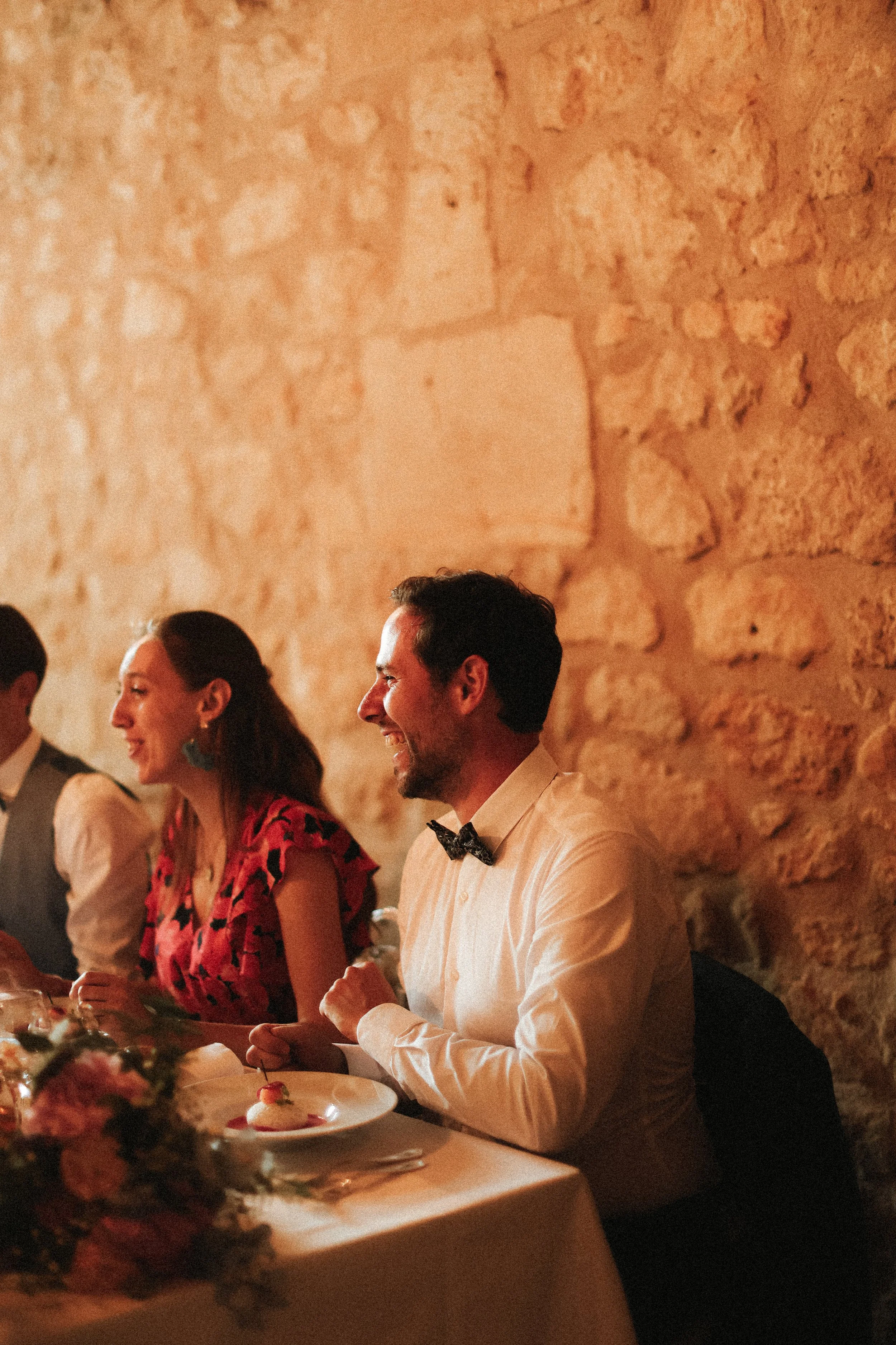 Des personnes souriantes assises à une table lors d'un dîner dans une salle avec un mur en pierre en arrière-plan.