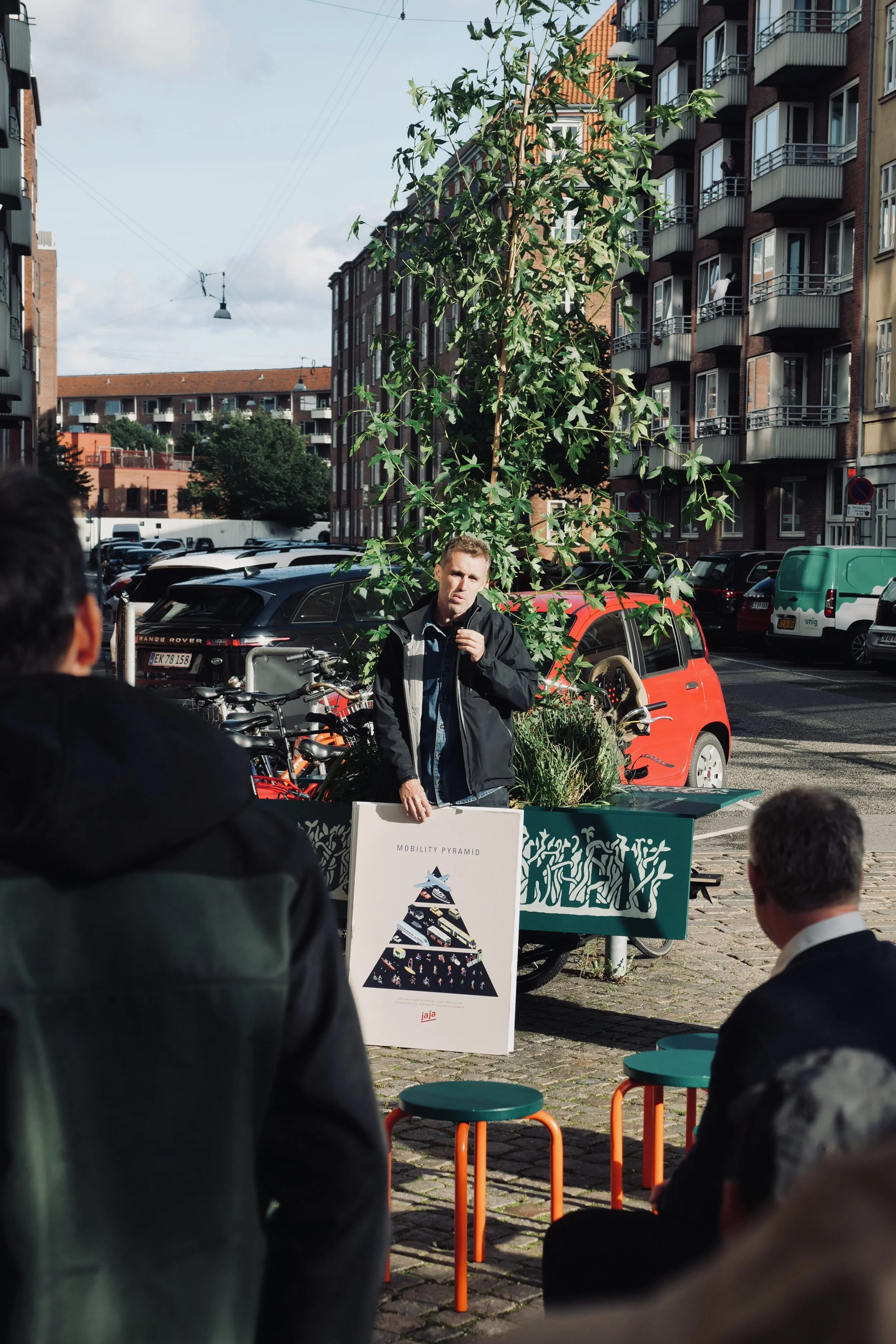 A man standing in front of a seated audience outdoors, delivering a presentation about a 'Mobility Pyramid' with a diagram on a sign, parked cars, and apartment buildings in the background.