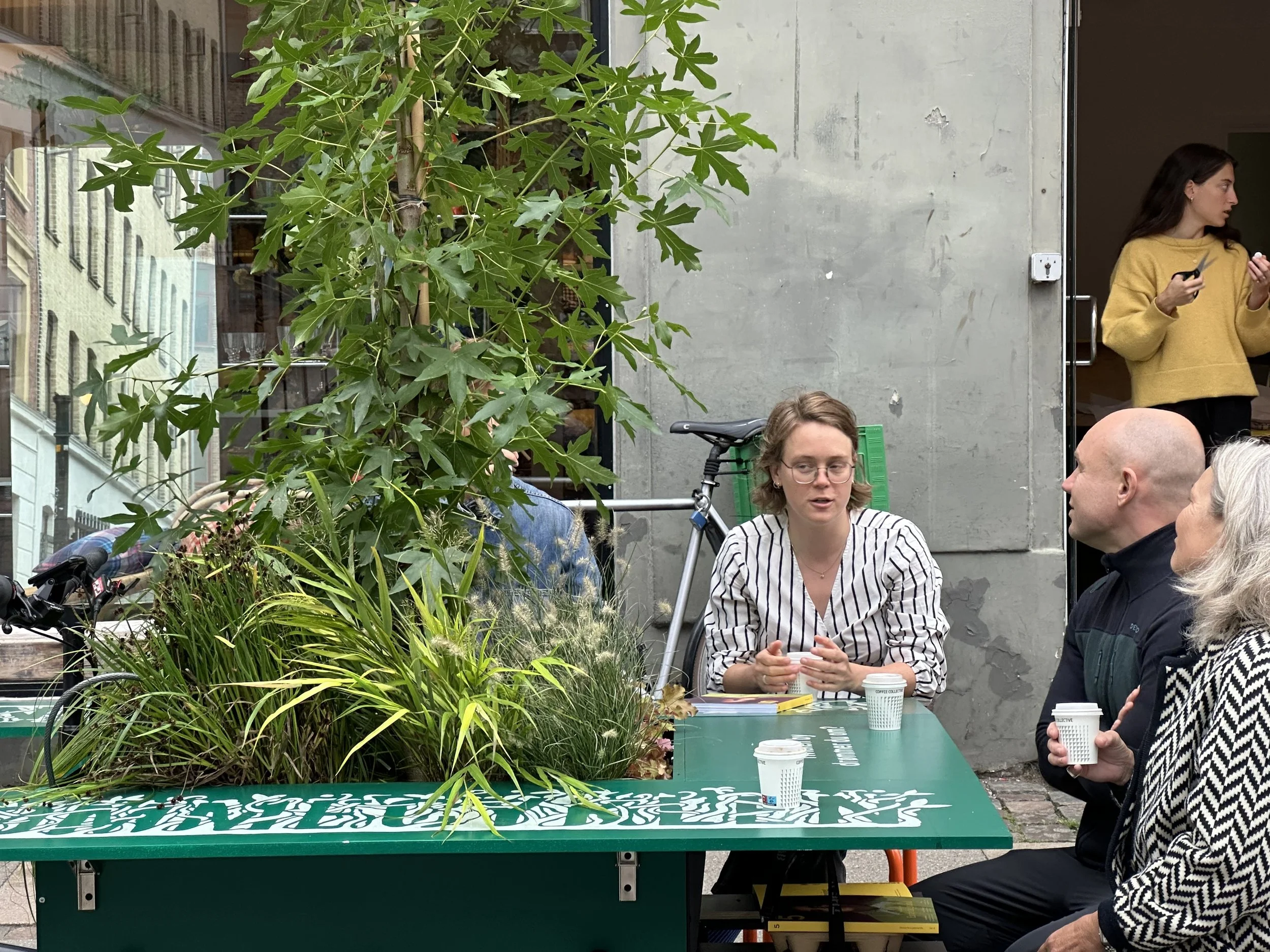 Three people sitting at a table outside, engaged in a conversation, with a large green plant and a bicycle behind them, and a young woman standing in the doorway holding a phone.