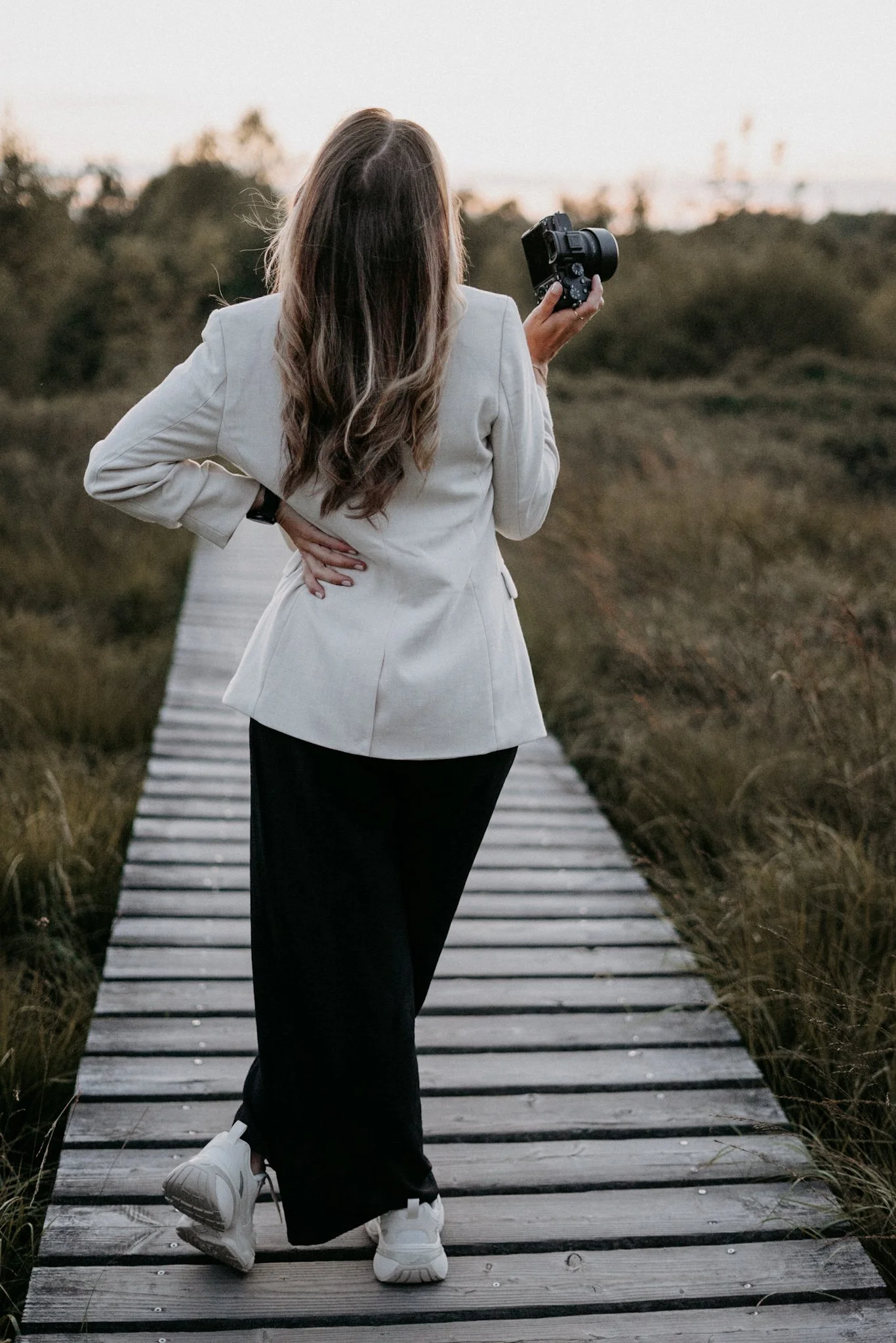Frau mit braunen, lockigen Haaren, in weißem Blazer und schwarzen Hosen läuft auf einem Holzsteg in der Natur, hält eine Kamera in der Hand.