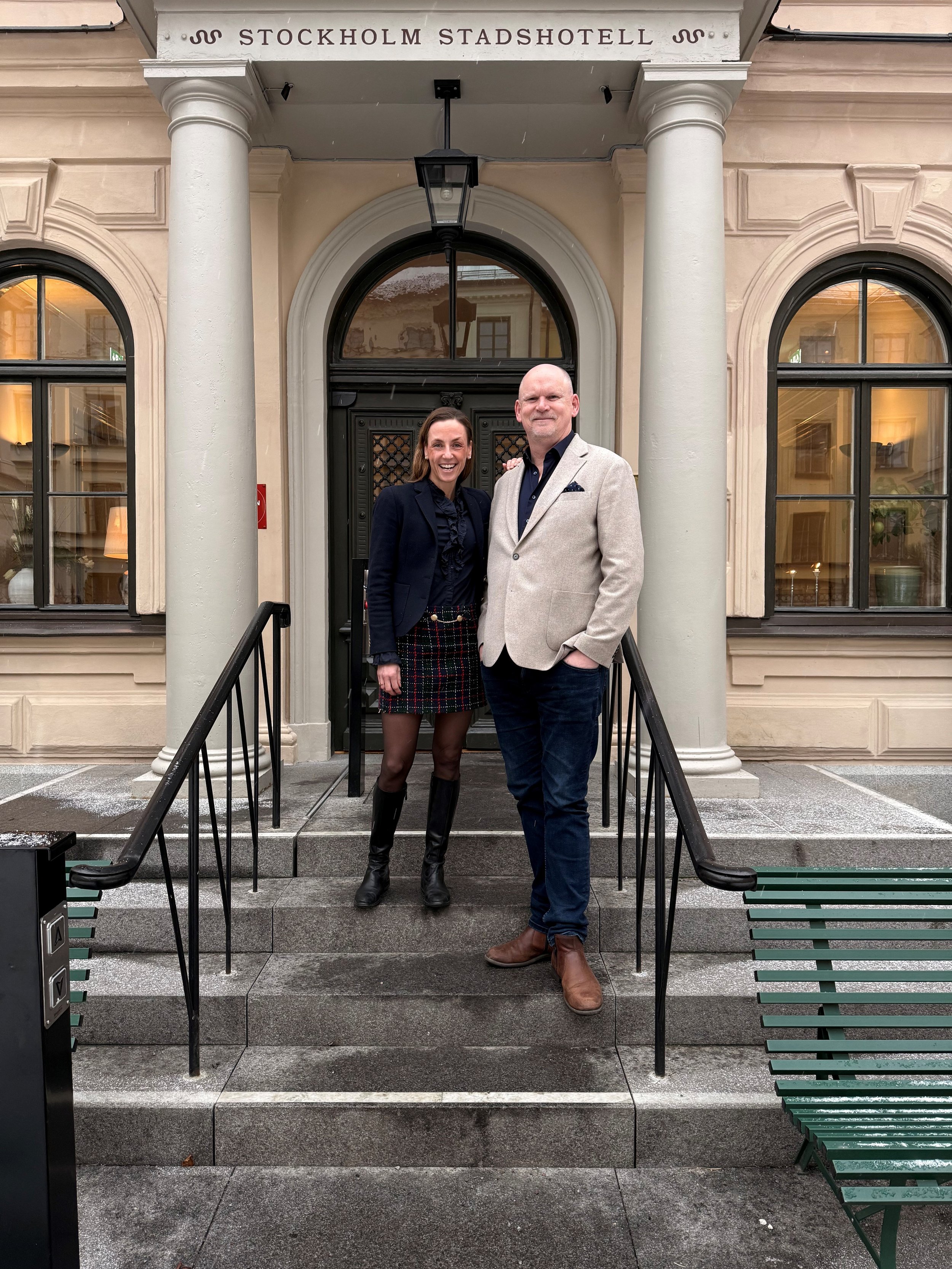 Två personer står på trappan framför ingången till Stockholm Stadshotell, ett kvinna och en man, båda ler. Kvinnan har mörkblå kavaj och mörka stövlar, mannen har beige blazer och bruna skor.