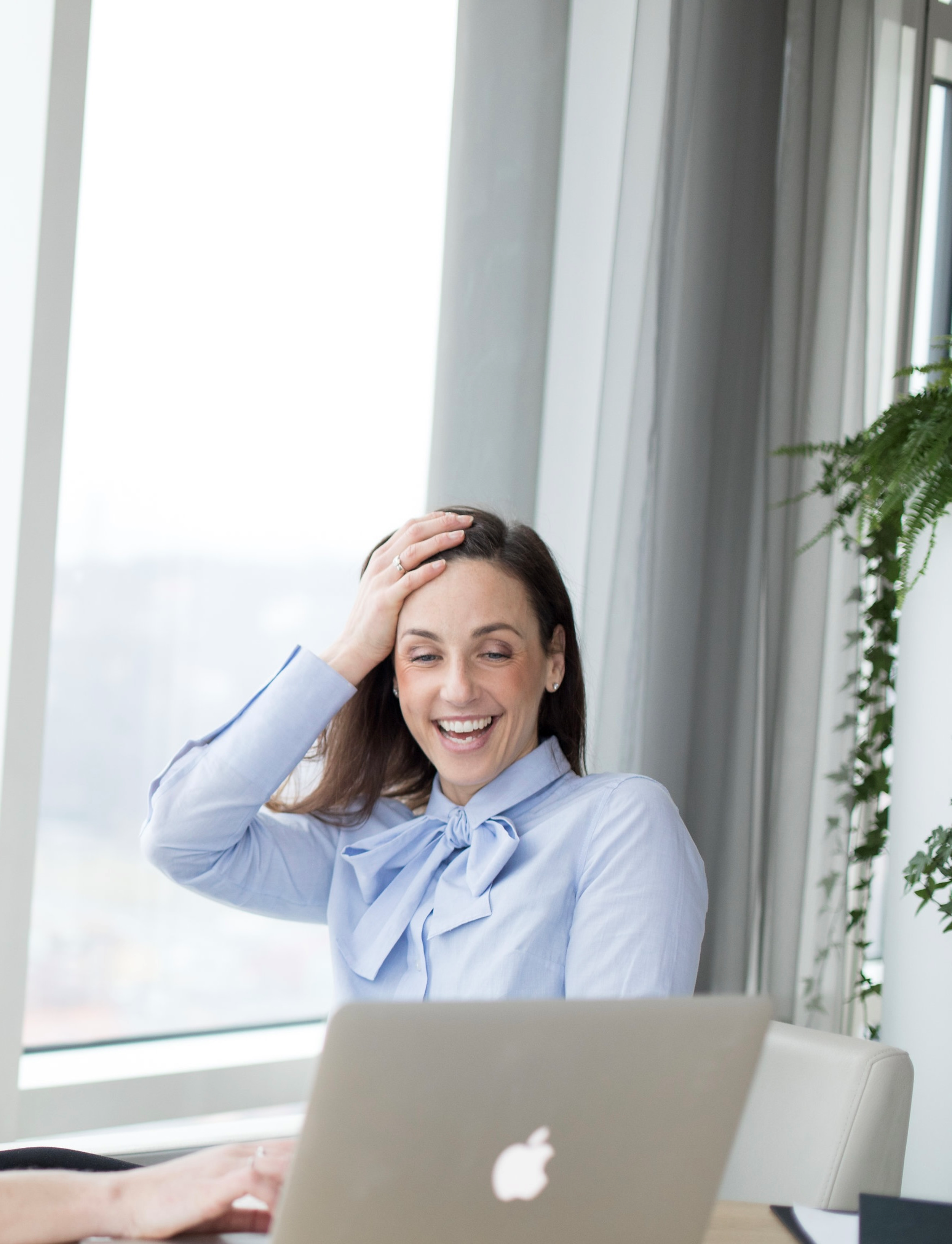 Kvinna i kontorsmiljö som ler och håller handen på huvudet, framför en laptop.