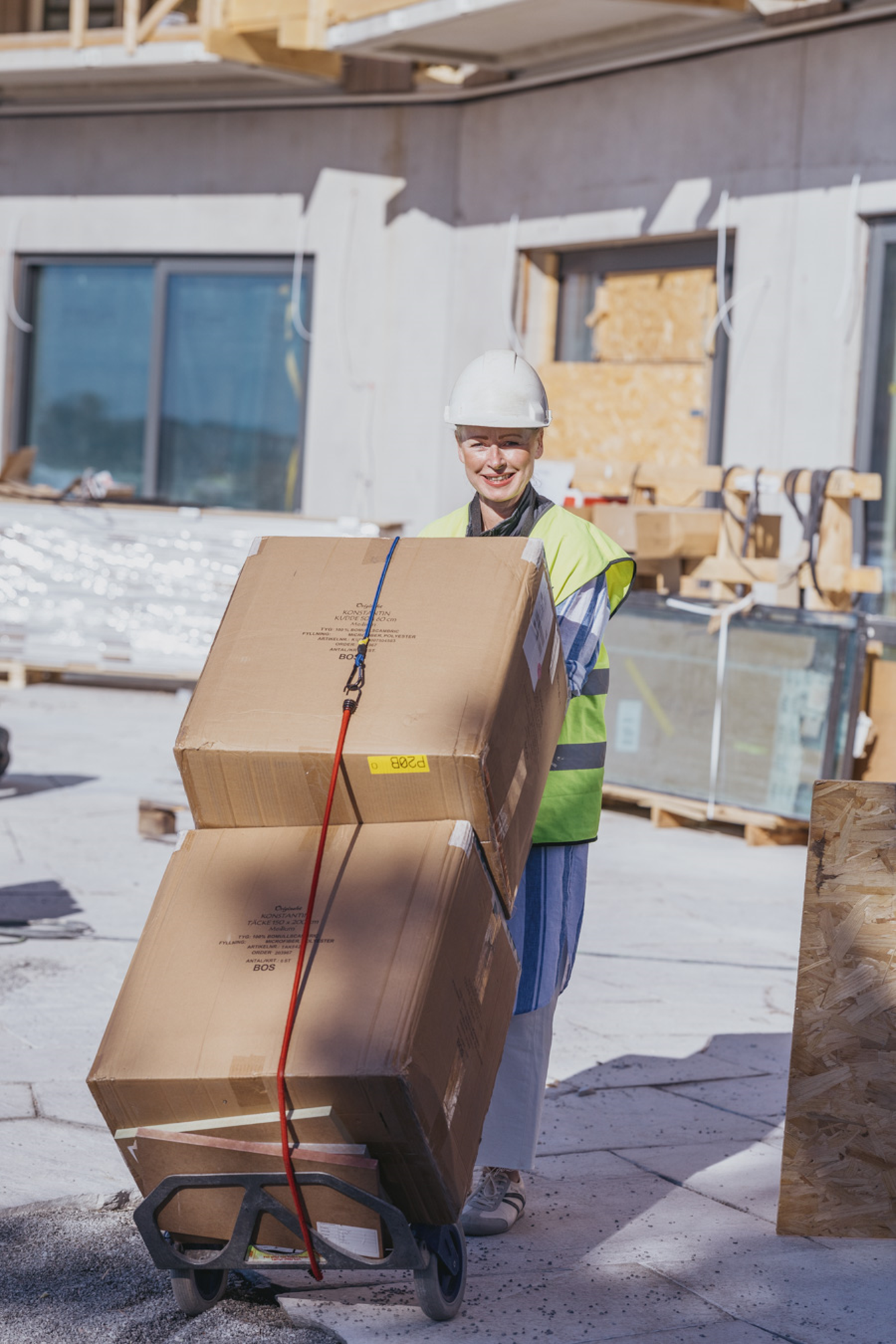 En kvinna i byggarbetskläder och vit hjälm som lyfter två stora kartonger på en handtruck på en byggarbetsplats.