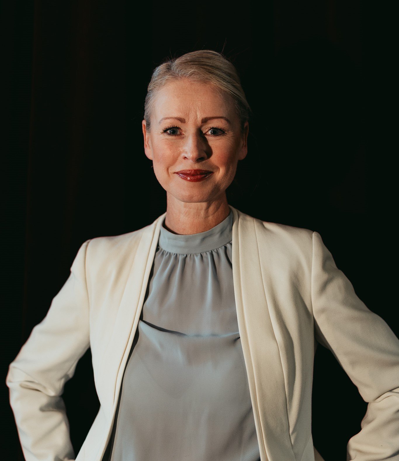 En kvinna med ljus hår och röd läppfärg, bär en vit kavaj och en ljus top, står framför en mörk bakgrund.
