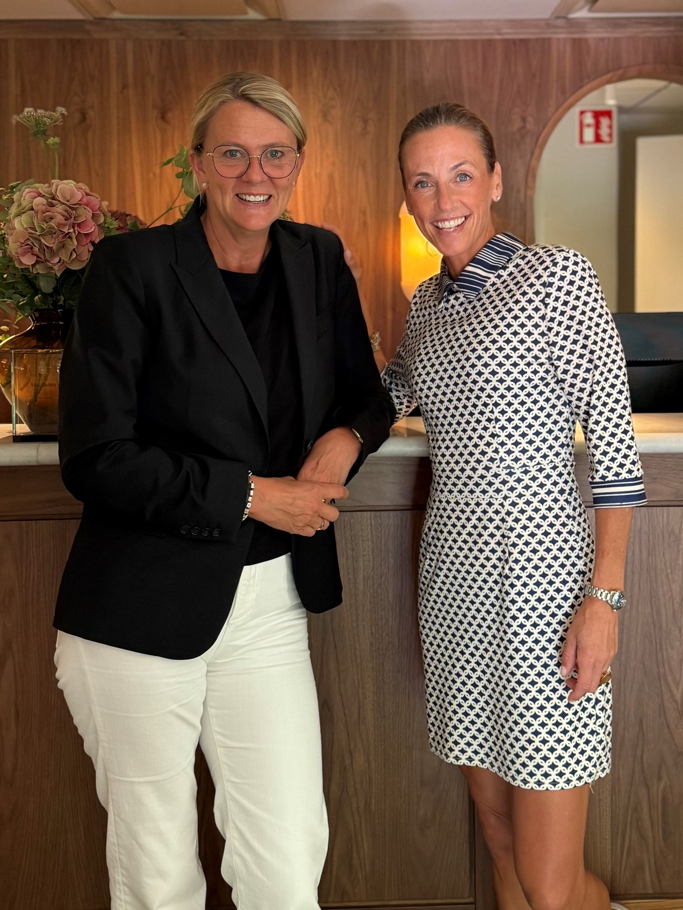Två kvinnor står bredvid varandra framför ett receptiondisk, båda ler mot kameran. En kvinna bär en svart kavaj och vita byxor, den andra har en mönstrad klänning.