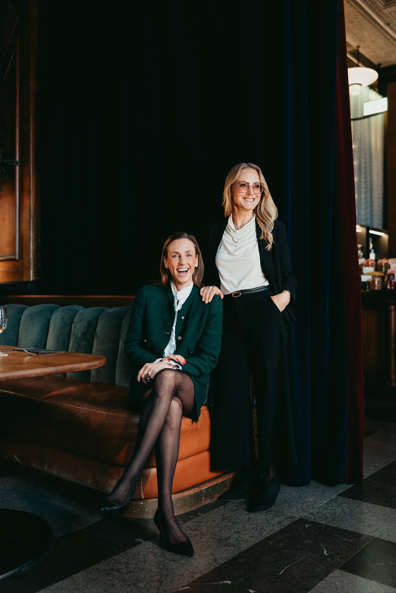 Två kvinnor i formella kläder poserar i ett elegant restaurang- eller barutrymme, en av kvinnor sitter och den andra står bredvid henne, båda ler glatt.