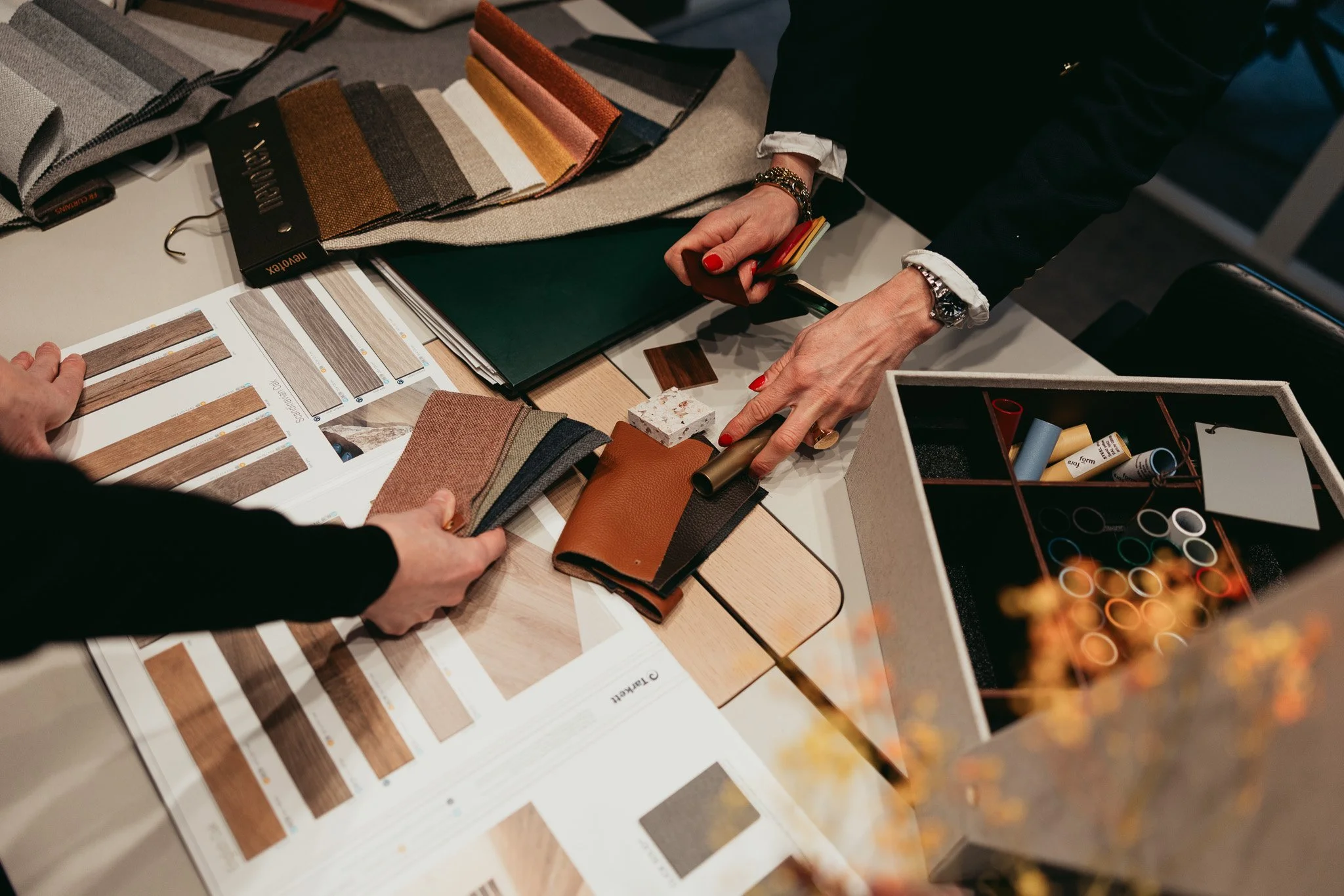 Två personer som jämför olika tyger och material för inredning, med färgprover, tygprover, och kataloger på ett bord.