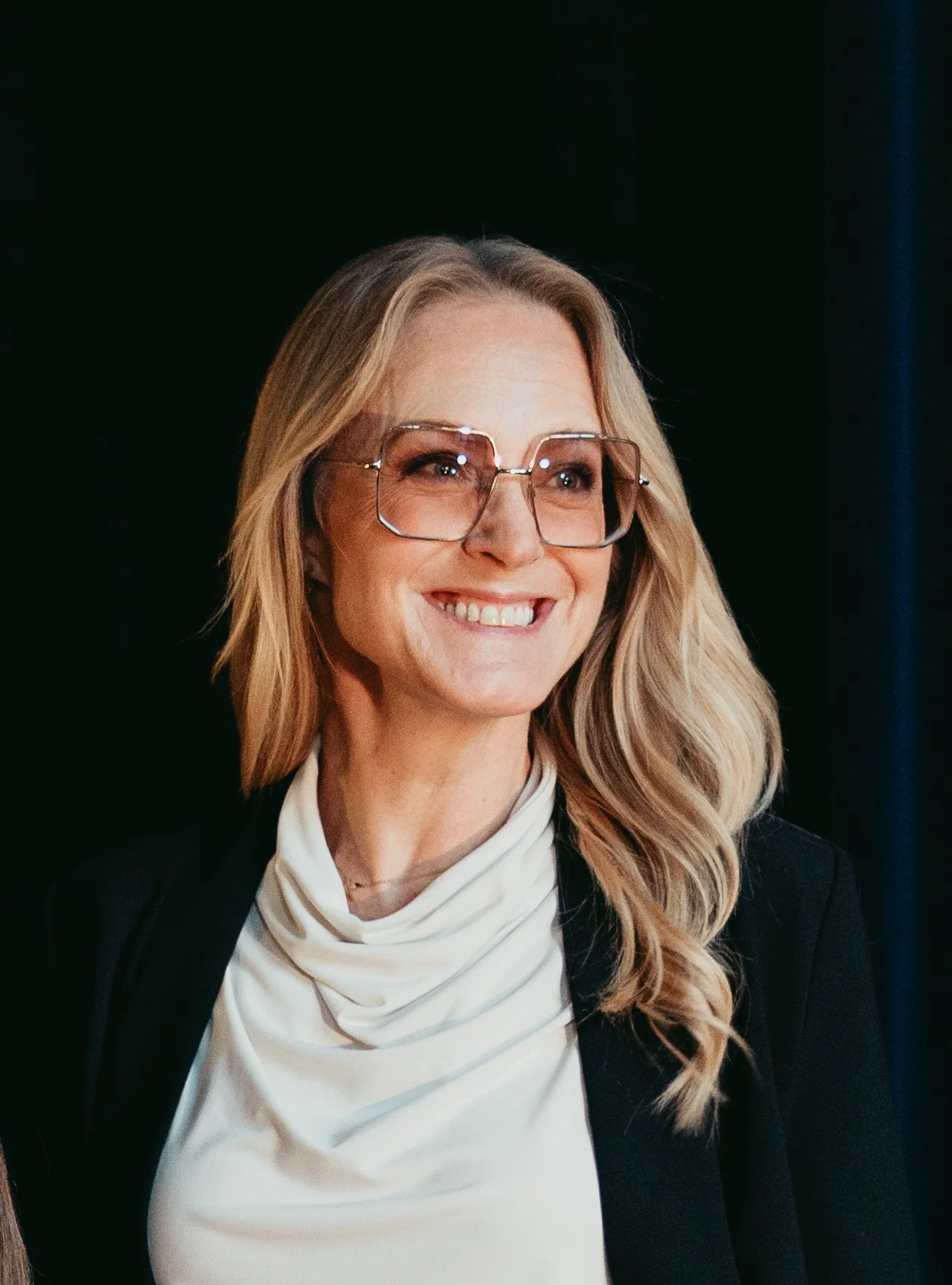 En leende kvinna med långt blont hår, stora glasögon, vit halsduk och svart jacka mot en mörk bakgrund.