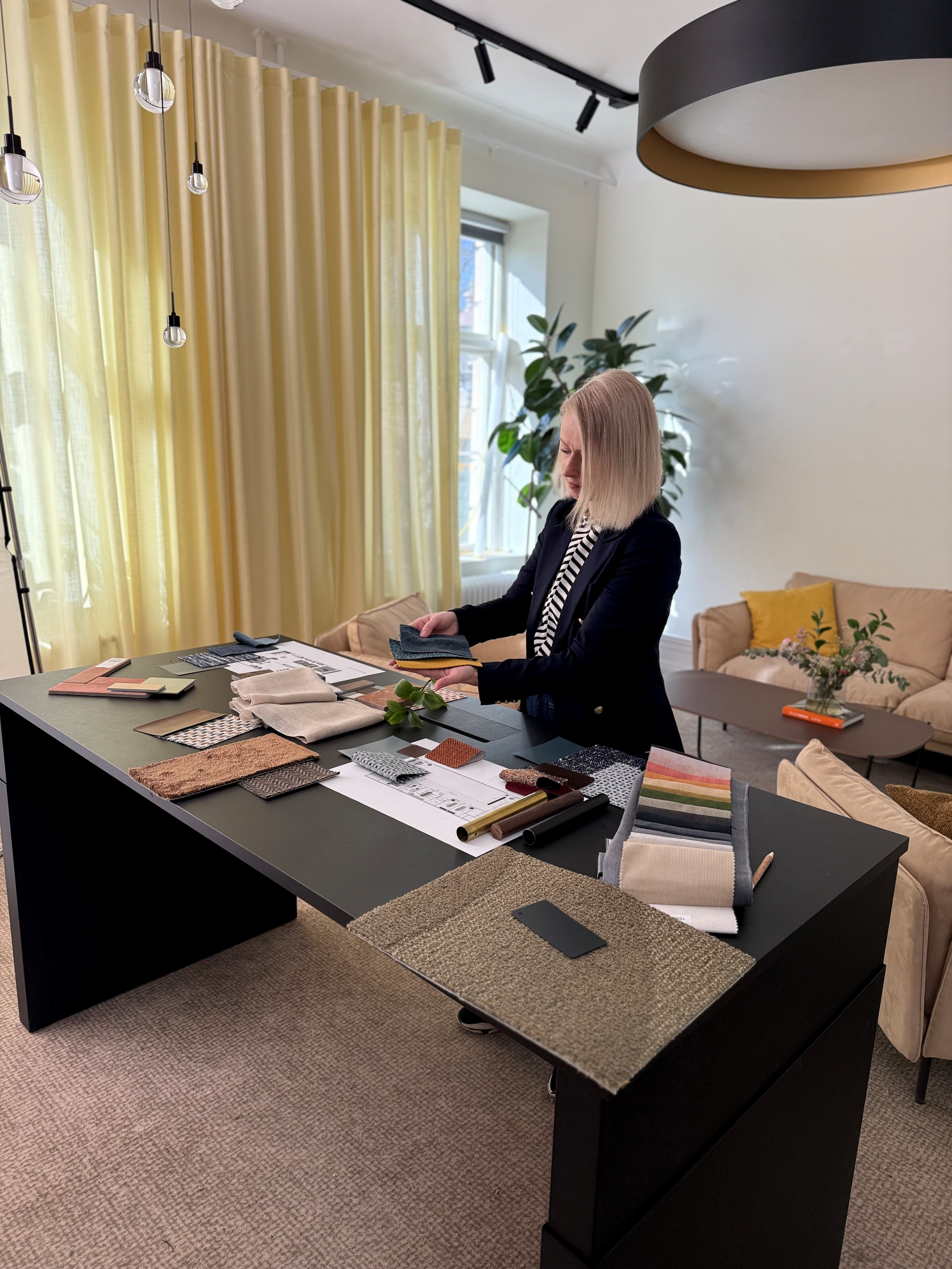 Kvinna som bläddrar i tygprover vid ett bord med olika mönster och färger, i ett inrett hem eller kontorsmiljö med gröna växter, ljusa gardiner och moderna lampor.
