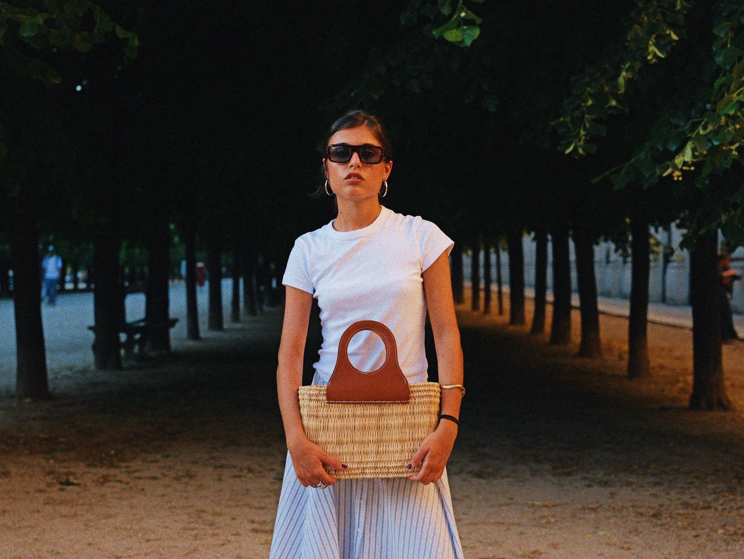 Jeune femme portant des lunettes de soleil, une robe blanche et tenant un sac en paille, se tenant dans un parc ombragé