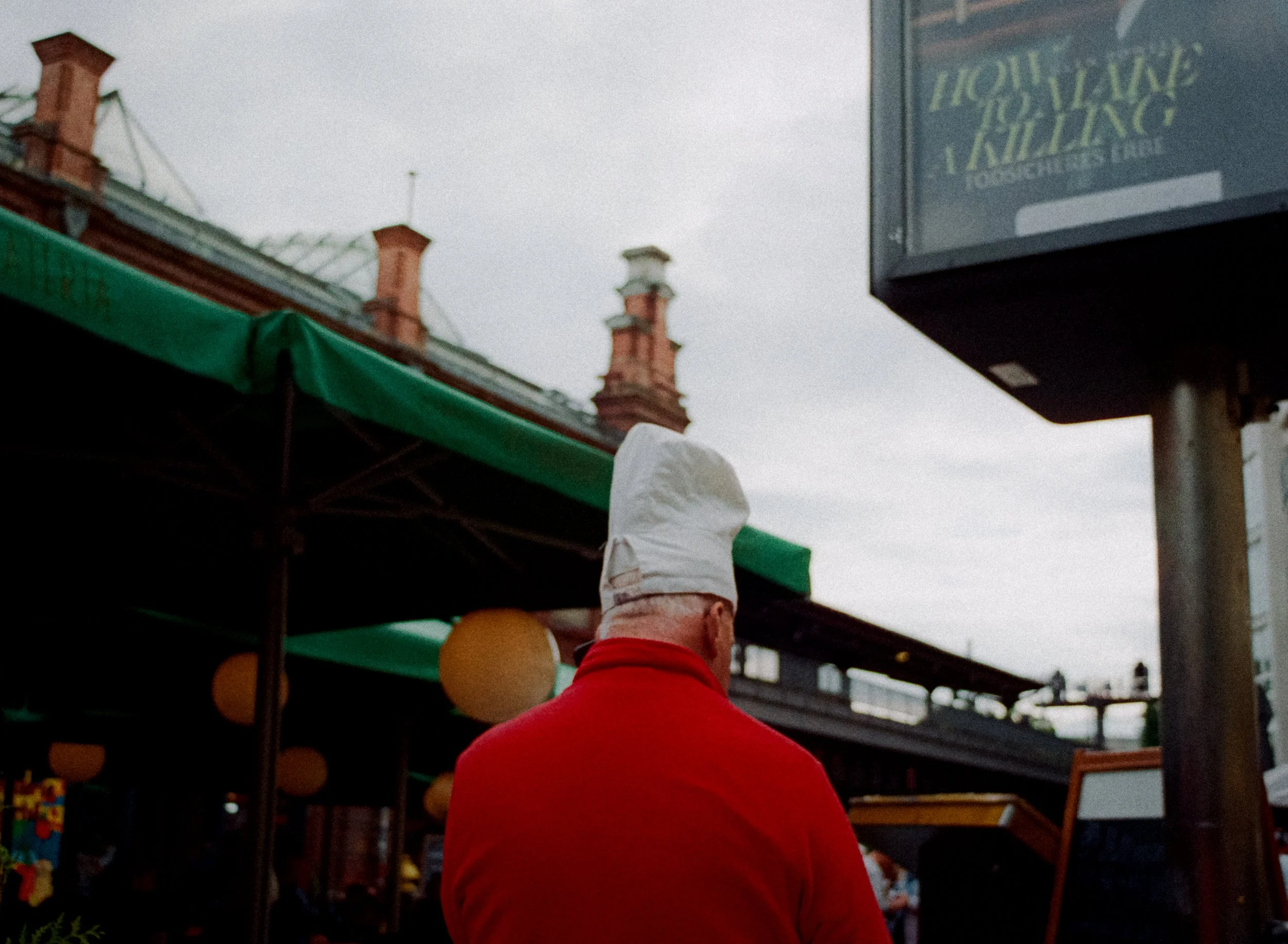Une personne avec un chapeau blanc vu de dos, portant un vêtement rouge, dans un marché extérieur sous un ciel en partie nuageux. Des structures en bois ou métal avec des toiles vertes sont visibles.