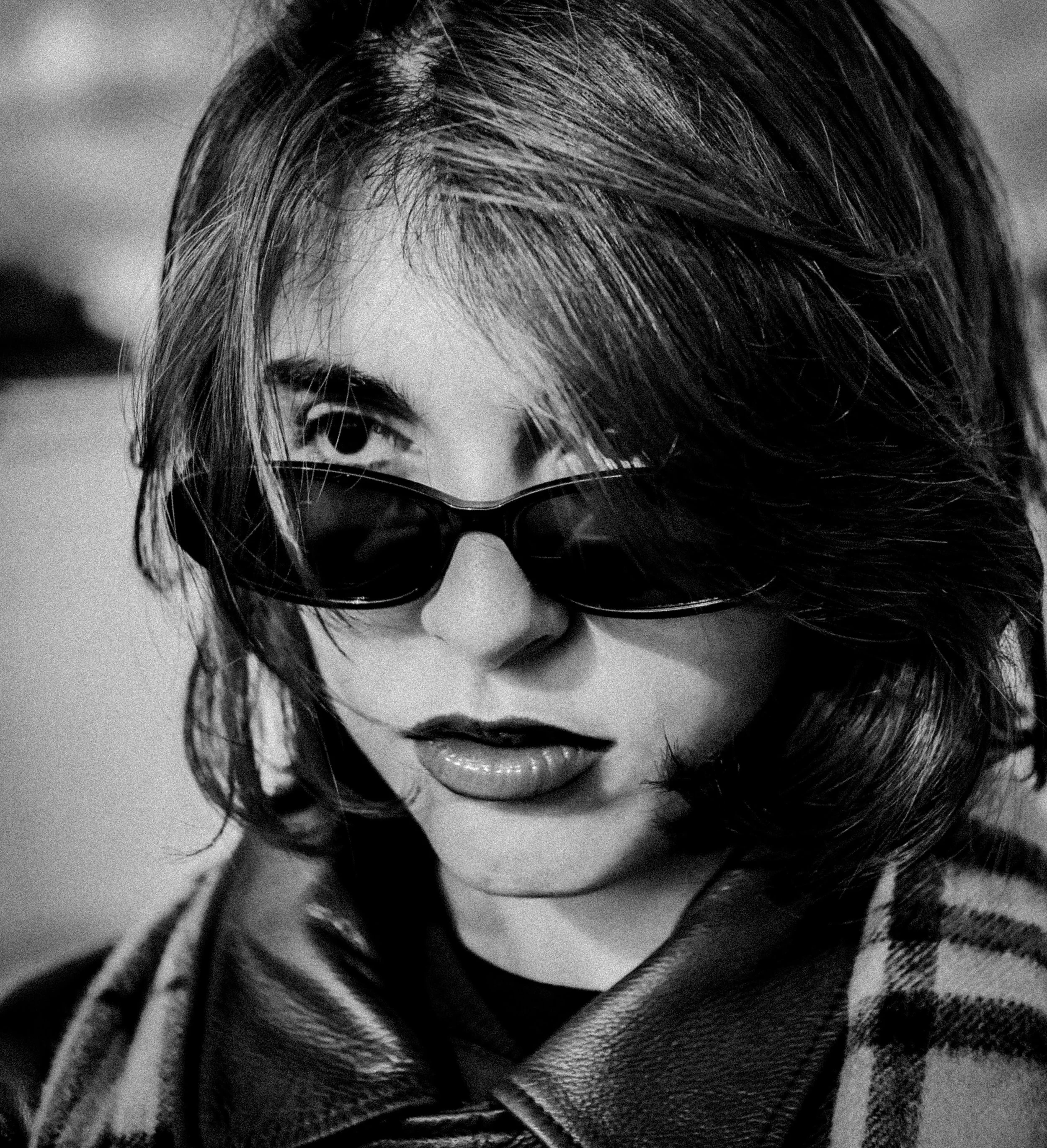 Un portrait en Noir et Blanc d'une femme portant des lunettes de soleil, avec des cheveux mi-longs et bouclés.
