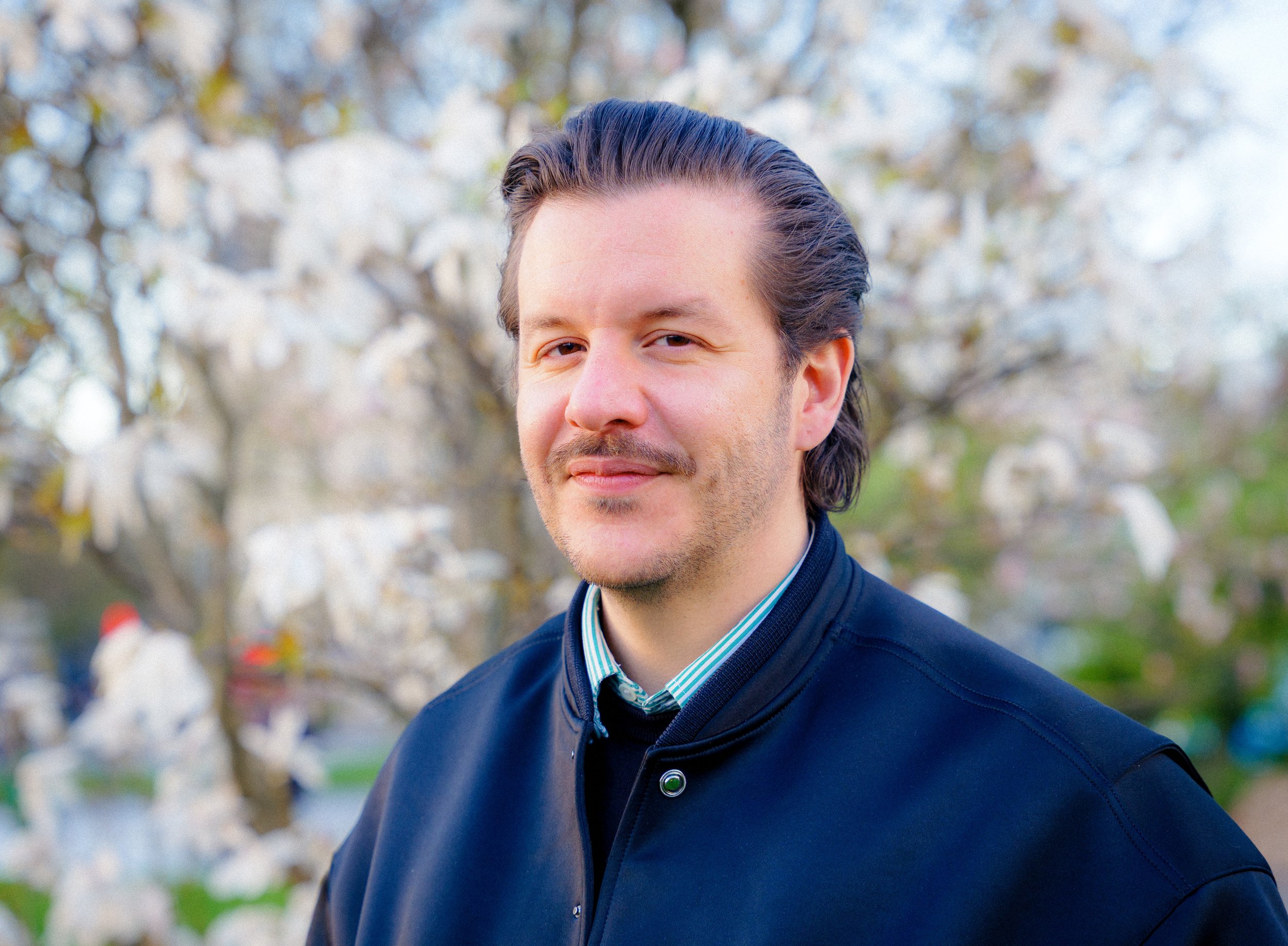 Portrait d'un homme avec des cheveux longs et foncés, portant une chemise à rayures et un vêtement foncé, se tenant à l'extérieur avec un fond de branches d'arbres en fleurs.