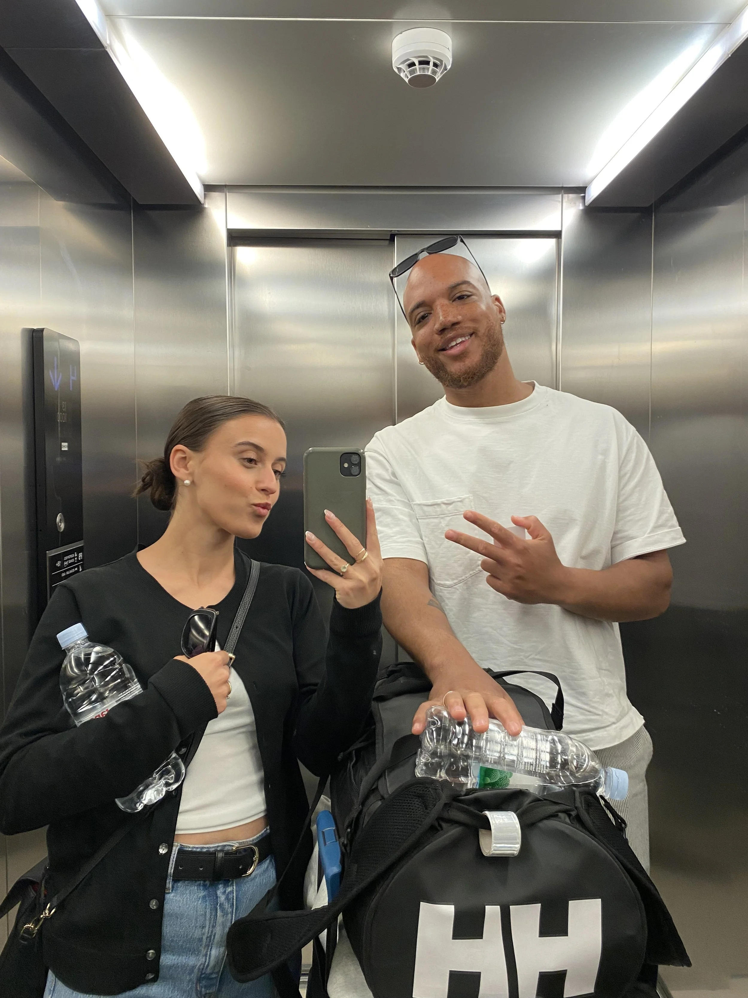 Two people taking a selfie in an elevator. The woman on the left is holding a water bottle, and the man on the right is pointing with his fingers. They are smiling and standing next to a black gym bag with water bottles.