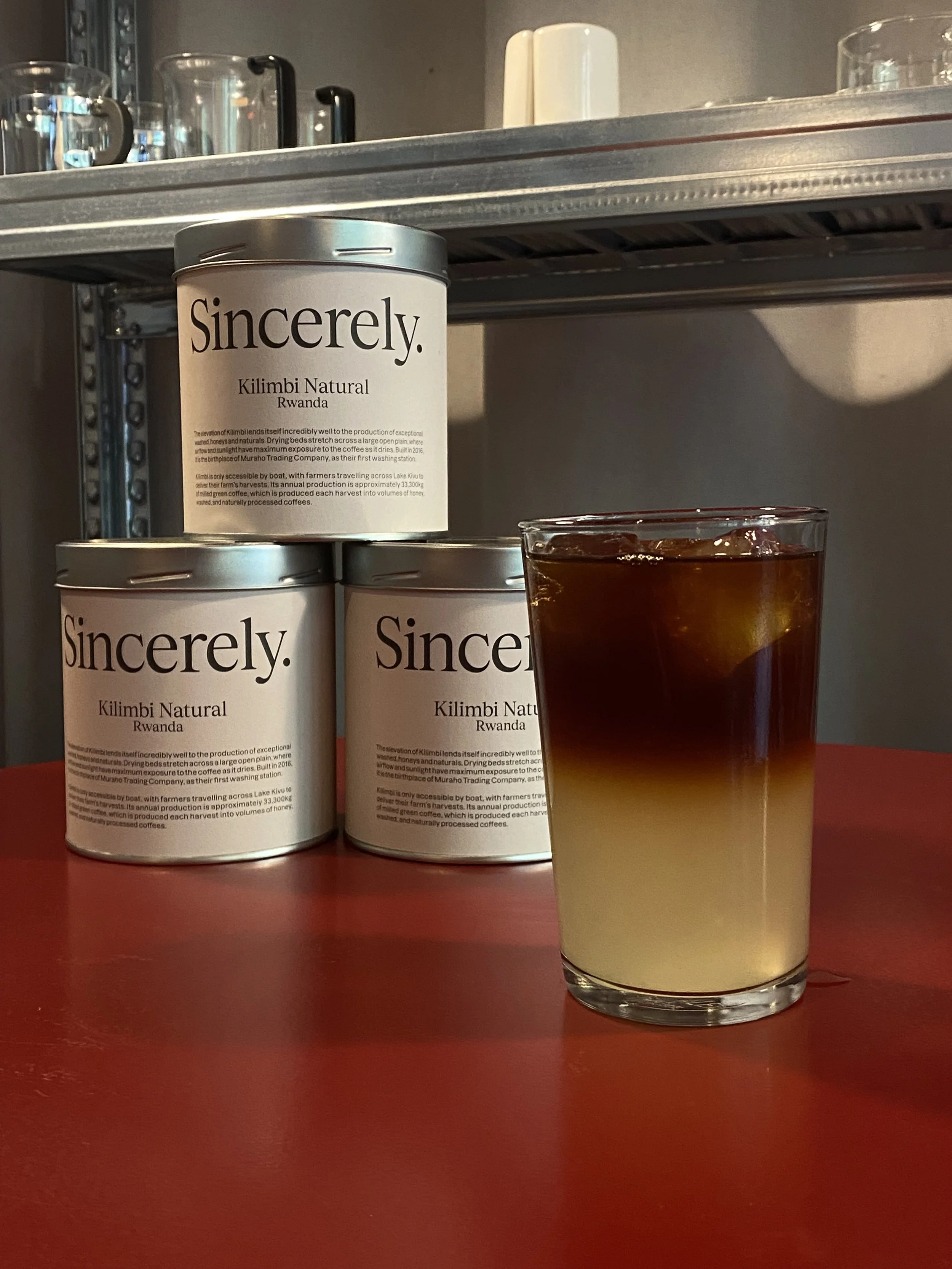 Three cans of Sincerely Kilimbi Natural Rwanda coffee on a red table, with a glass of iced coffee in front.