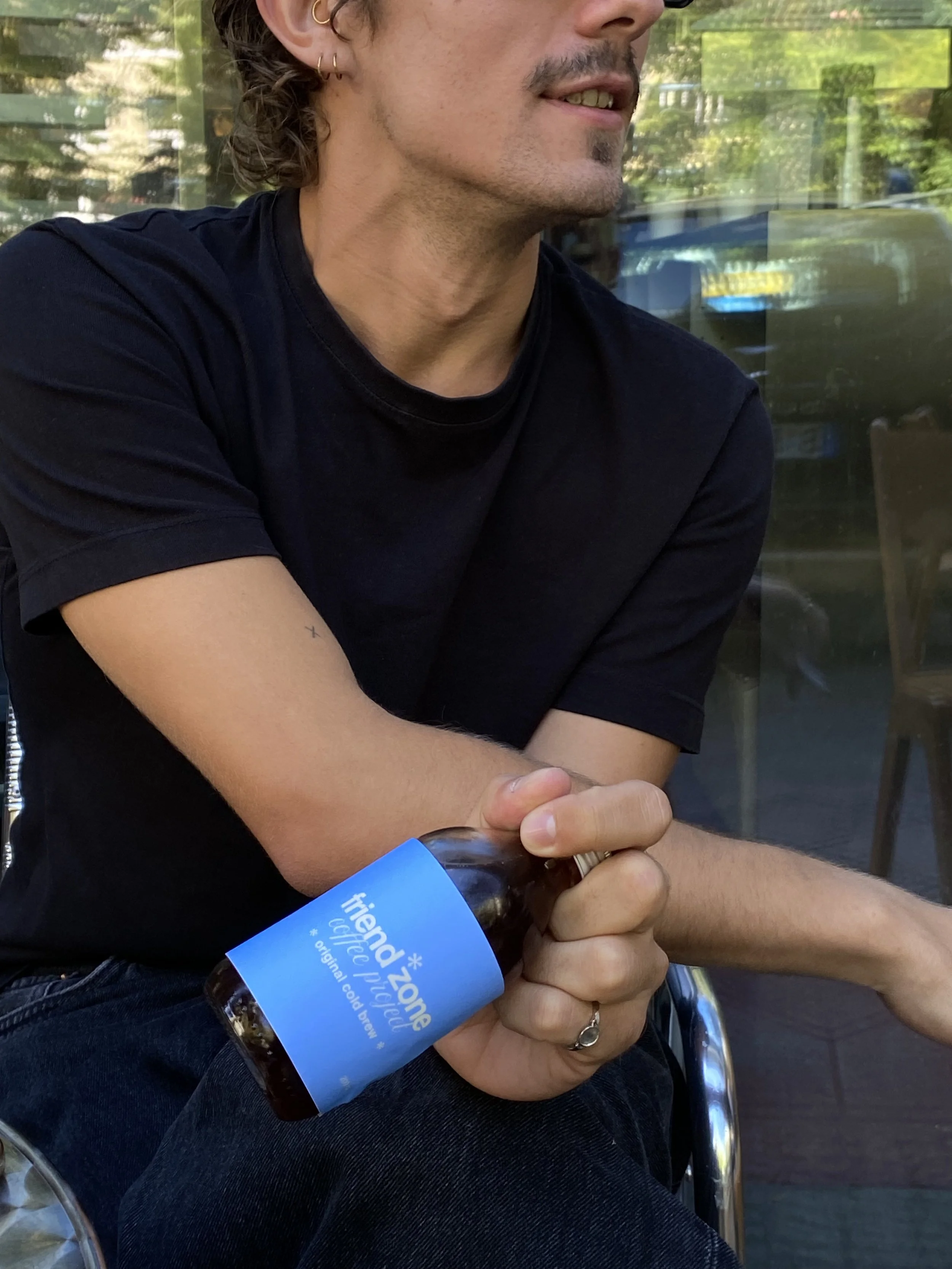 A person with light skin, facial hair, and earrings sitting outdoors, holding a brown glass bottle with a blue label that reads 'friend zone'. The person is wearing a black t-shirt and has tattoos on their arm.