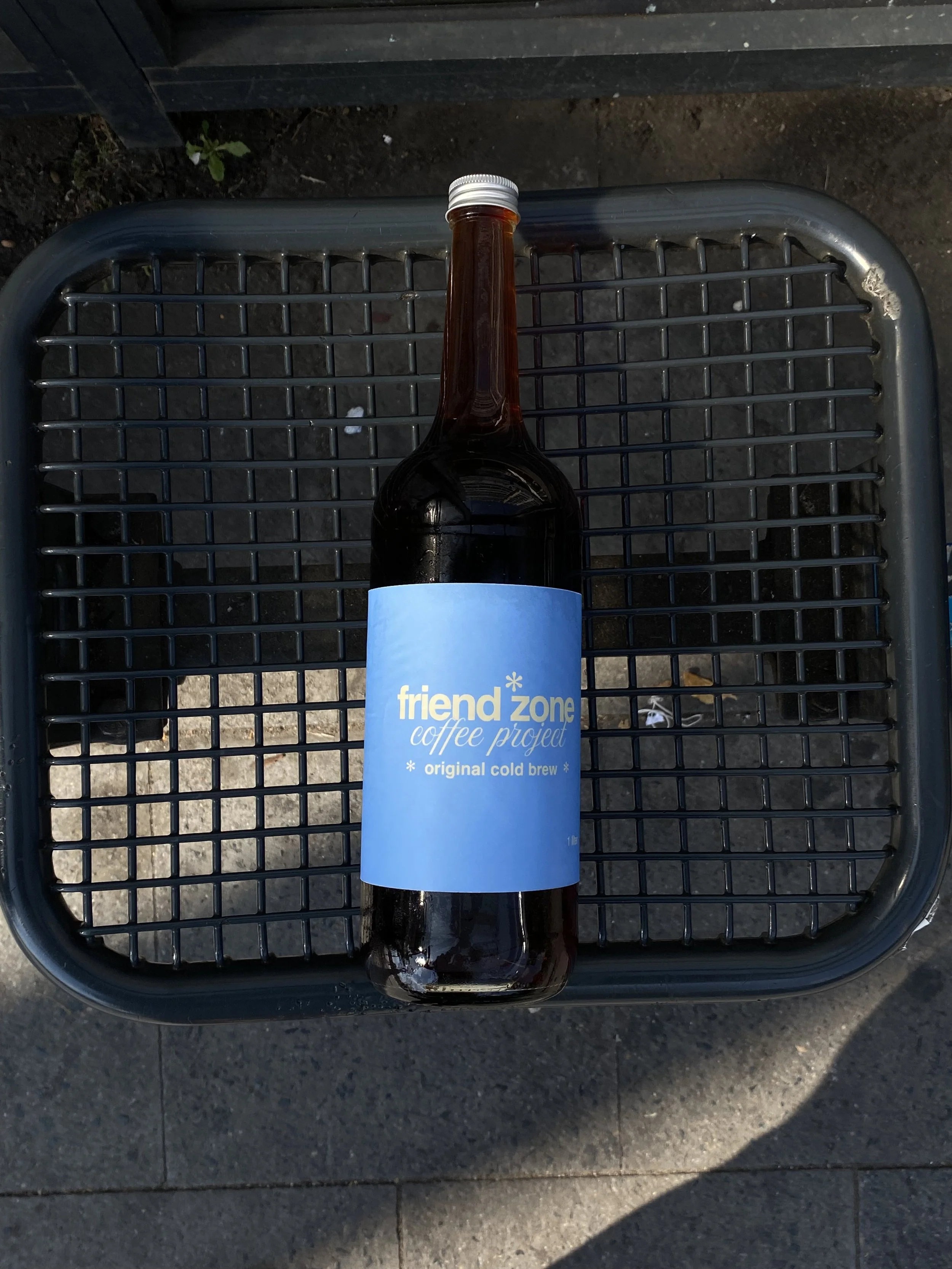 A brown glass bottle with a blue label reading 'friend zone coffee project, original cold brew,' placed on a wire mesh surface, outdoors.