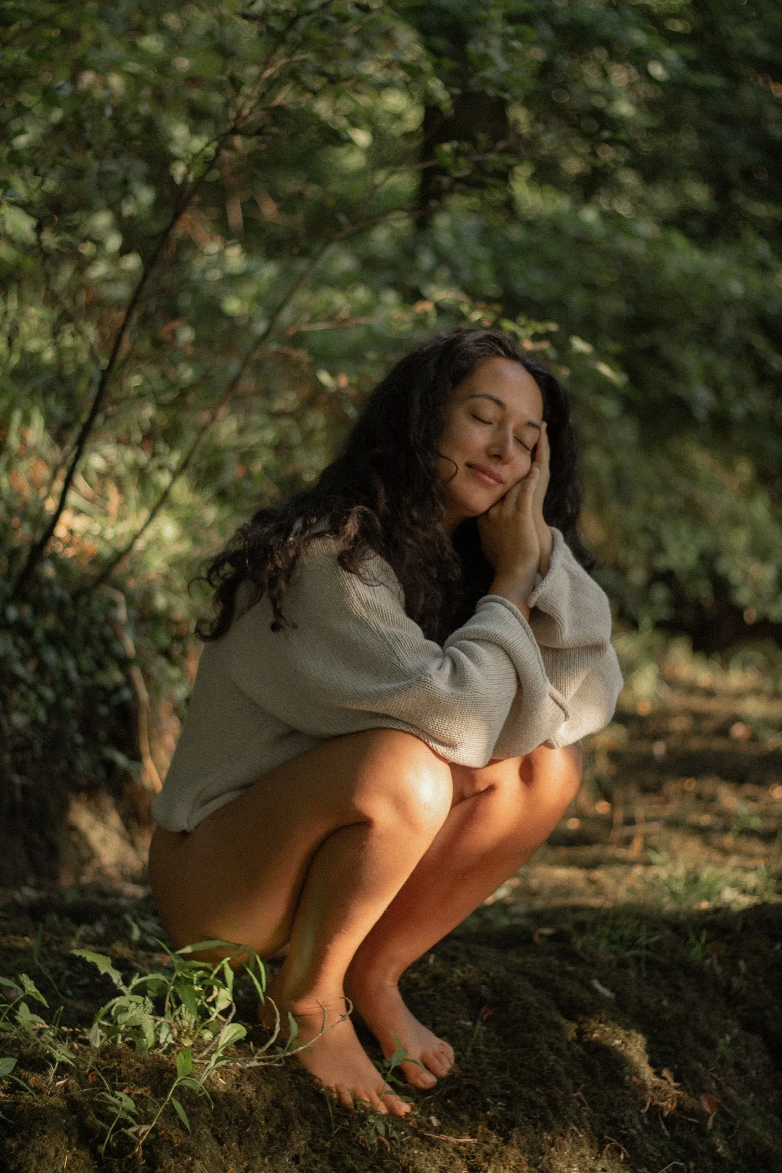 A woman squatting outdoors in a forest, with her eyes closed and her hand resting on her cheek, appearing relaxed and peaceful.