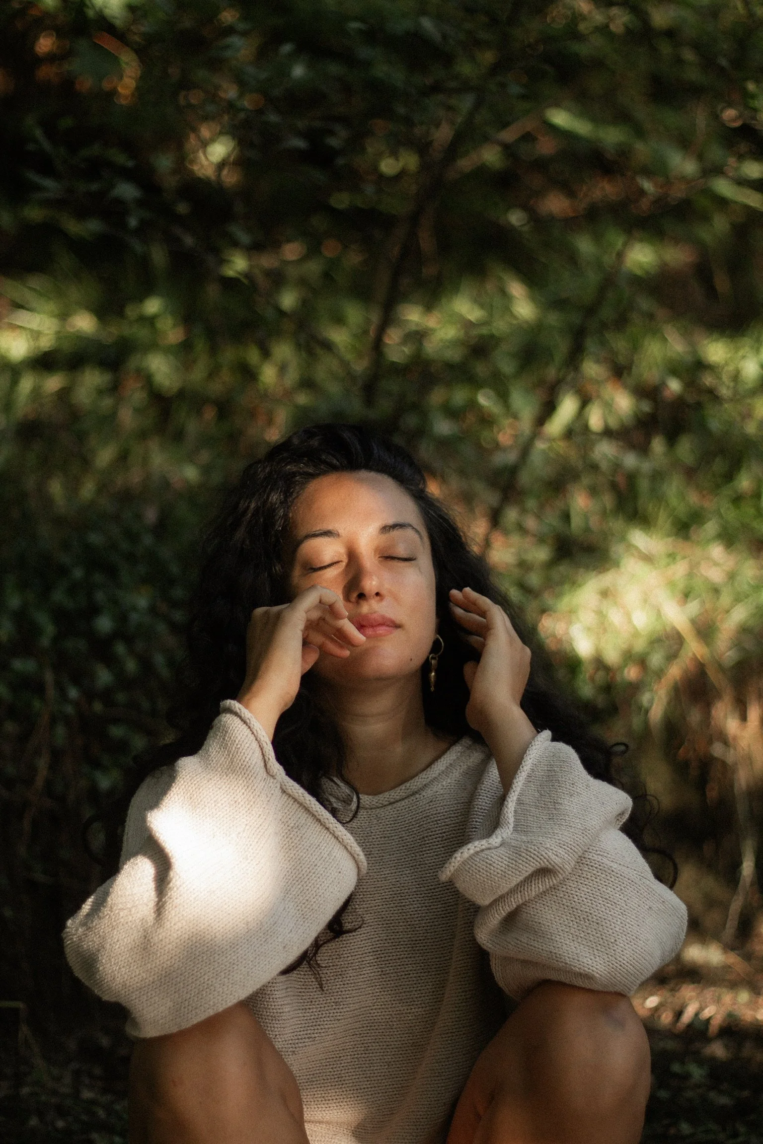 A young woman with long curly dark hair, wearing a beige sweater, sits outdoors with her eyes closed and a peaceful expression, surrounded by greenery and dappled sunlight.