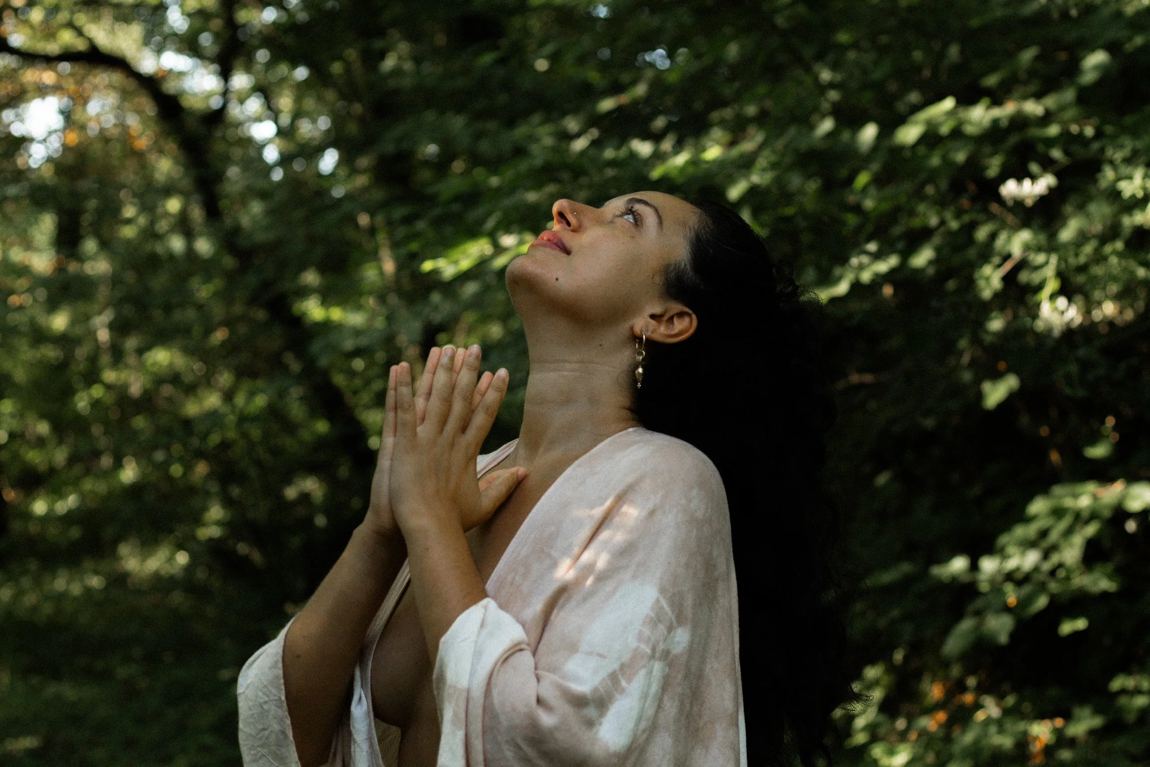 A woman standing in a forest with her hands held together near her chin, looking upward with her eyes closed, wearing earrings and a light-colored robe.