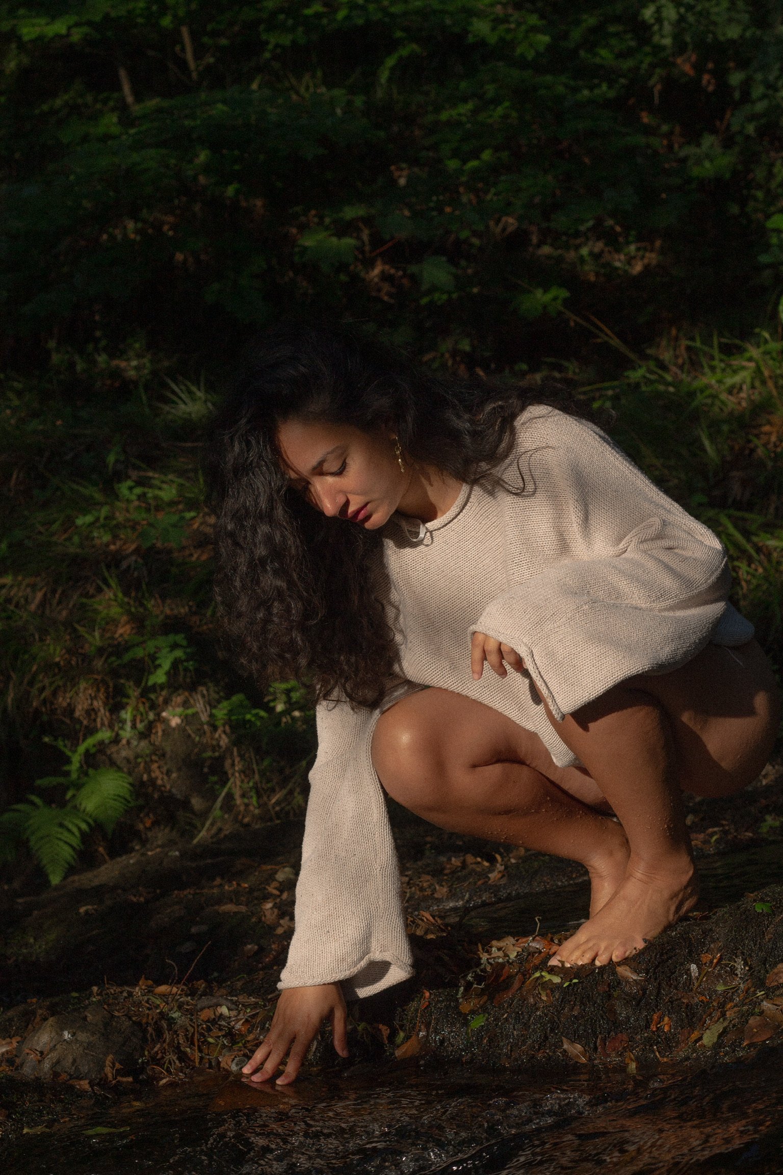 A woman with long curly hair crouches by a stream in a forest, touching the water with her right hand. She is wearing a beige oversized sweater and appears to be in a peaceful, contemplative mood.